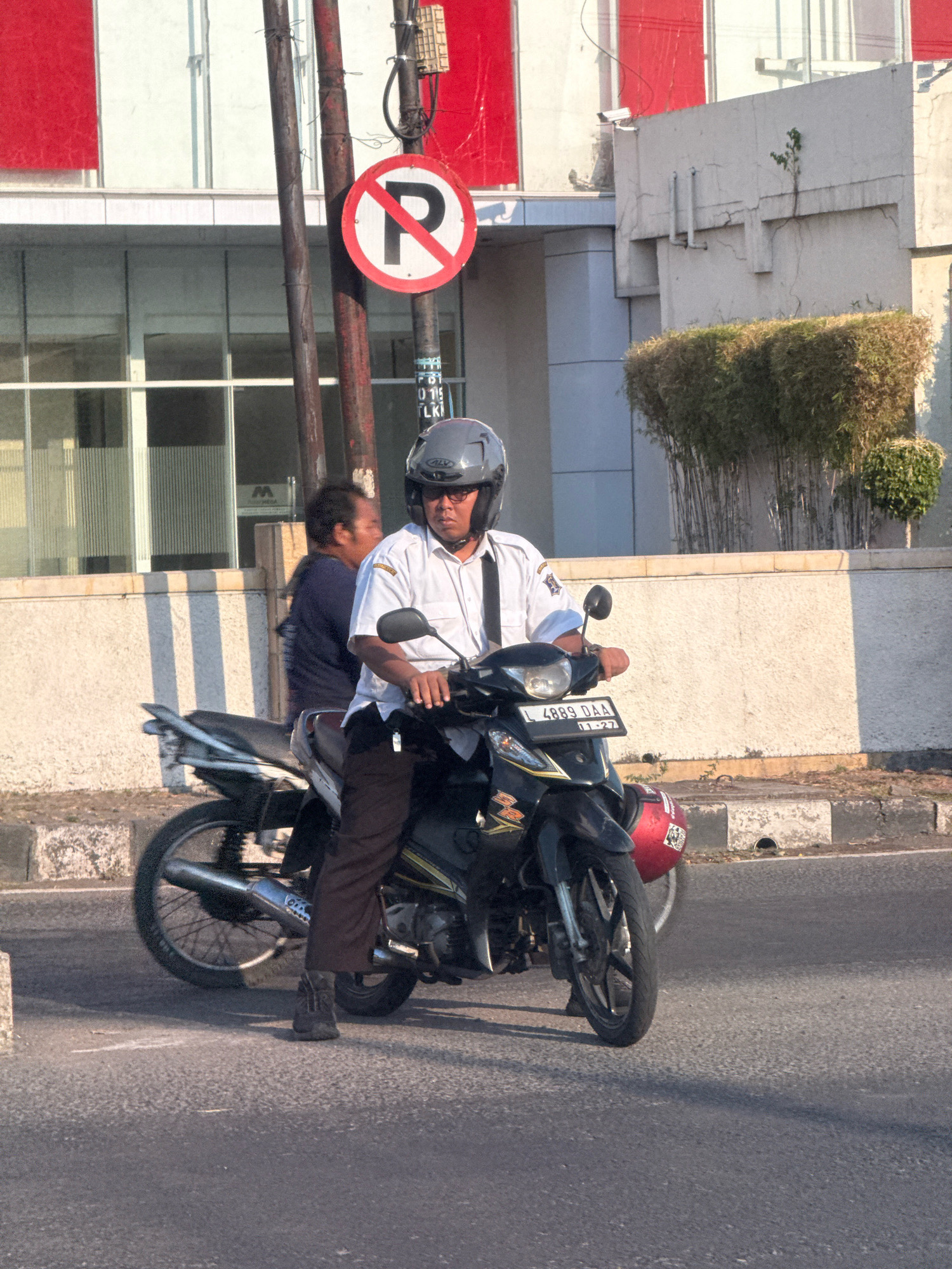 An Indonesian man wearing a white collared shirt, dark pants, and a gray helmet rides a black motorcycle on a paved street in Surabaya, Indonesia. The motorcycle has a license plate that reads "L 4889 DAA". He is wearing sunglasses and appears to be looking forward. Behind him, another man in a dark shirt stands near a utility pole. A "No Parking" sign is visible on the utility pole. The scene is illuminated by sunlight, casting shadows on the road. In the background, there is a modern building with red accents and a line of green plants. The street has a concrete curb and gutter.