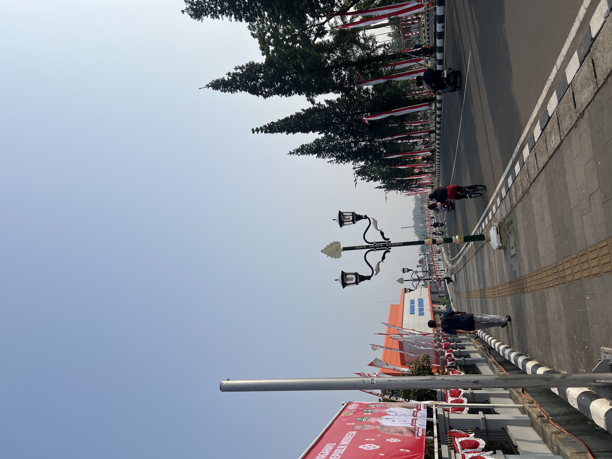 The image depicts a street scene in Cibinong, Indonesia, likely near the Cibinong City Mall. The street is lined with tall, slender pine trees and decorated with numerous Indonesian flags. The flags are red and white, alternating with ornamental white fabric banners. A prominent banner on the left side of the image, red with white text and images of people in uniform, reads "DIRGAHAYU REPUBLIK INDONESIA."
On the sidewalk to the left, a single person walks away from the camera, wearing a dark t-shirt and light-colored pants. To the right of the sidewalk, a road is visible with traffic. Two individuals are riding bicycles, and two others are on motorcycles. A distinctive street lamp with an ornate, gilded finial stands on the sidewalk. In the background, a building with an orange facade and white trim is visible, with the text "UPTD PERPUSTAKAAN KOT..." inscribed on it, suggesting it is a library. The sky is clear and bright blue, indicating a sunny day.