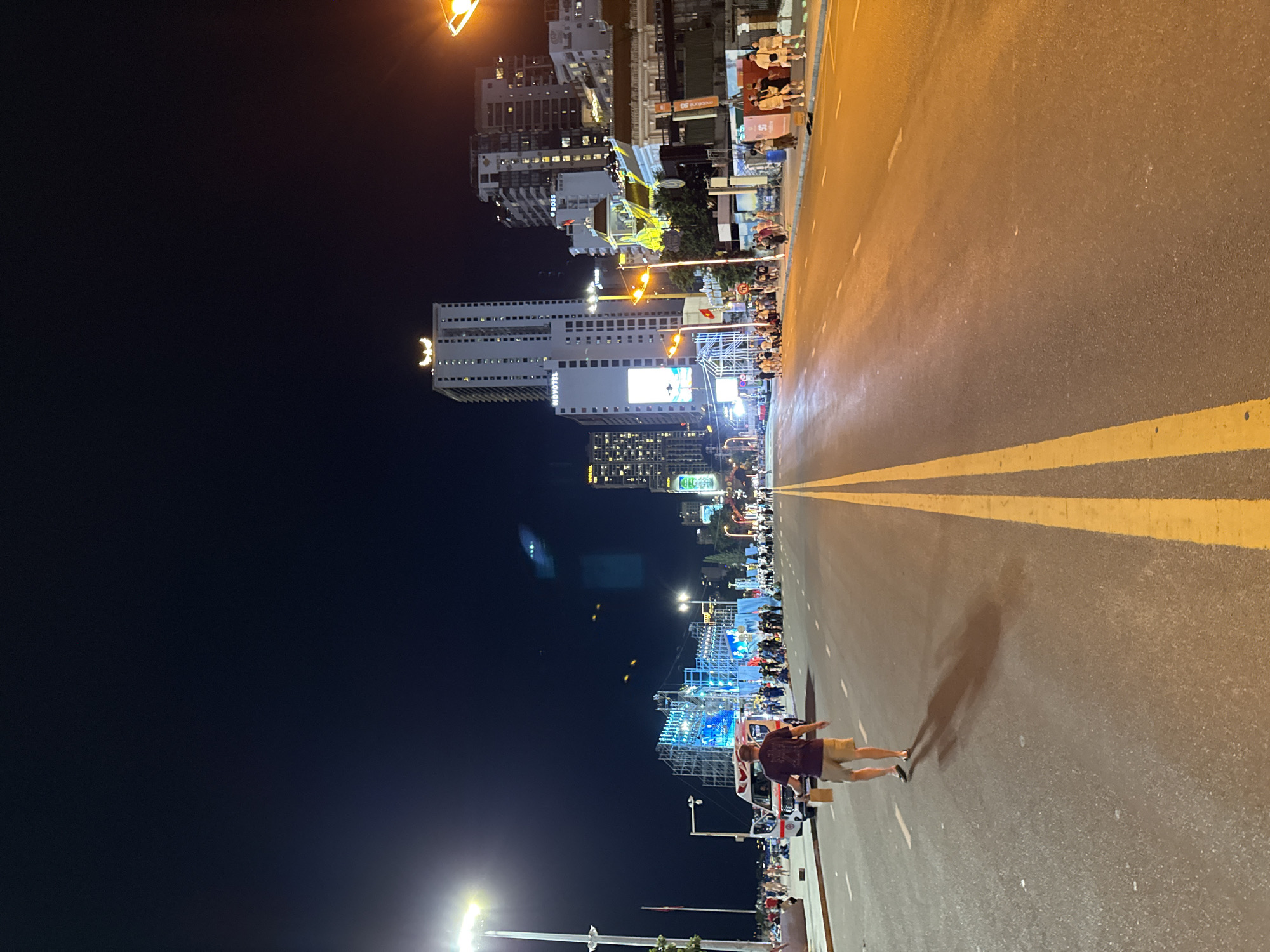 An eye-level, wide-angle nighttime shot captures a wide, empty street in Nha Trang, Vietnam, likely near the coast, illuminated by streetlights and building lights. The road is paved and has a yellow double line down the center. On the left side of the road, a man in a purple t-shirt and khaki shorts walks away from the camera, carrying a brown paper bag. Behind him, an ambulance with red and white markings is parked, and a crowd of people are gathered near what appears to be a stage or event setup with scaffolding and blue lighting. Buildings, some with illuminated signs like "NOVO TEL" and "BOSS," line the right side of the street. The sky is dark, with a few bright lights visible from street lamps and buildings, and some faint yellow and blue lights that may be part of a display or event further down the road. The atmosphere suggests a lively evening event or festival.