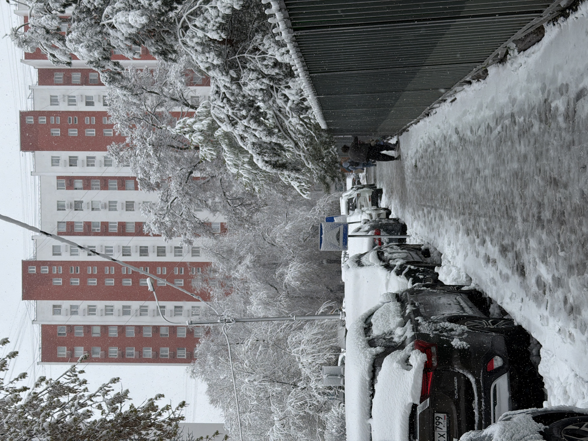 A wintry street scene in Moscow, Russia is depicted. Heavy snowfall blankets the sidewalk, parked cars, and trees lining the street. A tall apartment building with a red and white facade is visible in the background, indicating a residential area. Two people are walking on the snow-covered sidewalk. The cars are heavily covered in snow. The license plate on the visible car reads "KX 799 RUS".