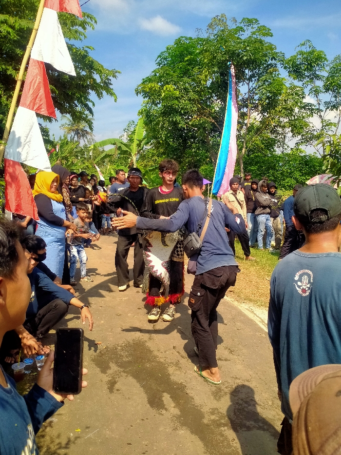 The image depicts an outdoor cultural event on a paved road in Plosorejo, Indonesia, under a clear blue sky with scattered white clouds. A crowd of approximately 20-30 individuals observes two central male figures.
The primary central figure, positioned slightly left of center and facing left, wears a dark long-sleeve shirt over a red undershirt with "SEMINAH EDAN" text, dark pants, and light-colored shoes adorned with yellow and red tassels. He also wears sunglasses, has dark curly hair, and is adorned with a white, black, and red traditional skirt-like garment featuring an abstract pattern around his waist. His arms are extended outwards. Directly in front of him, interacting, is another male figure, facing the primary figure with extended arms. This individual wears a dark blue long-sleeve shirt, dark cargo pants, a black shoulder bag, and green flip-flops.
To the left of the scene, red and white Indonesian flags are vertically attached to a bamboo pole. Among the spectators on the left, a woman in a yellow hijab and light blue top is visible, along with a young boy in blue jeans. In the immediate foreground on the left, a male in a blue shirt holds a black smartphone vertically, appearing to record the event. Small plastic cups are visible near him on the ground.
On the right side of the road, a tall, multi-colored (blue, pink, white) fabric banner is draped from a large, leafy green tree. Additional spectators, predominantly male, stand along the road's edge. One male in the extreme foreground right has his back to the camera, wearing a dark blue long-sleeve shirt with a white graphic logo (including a skull-like image and text such as "PSHT 1922") and a brown hat. The environment is characterized by lush green trees, banana plants, and vegetation bordering the paved road, which shows some dark, wet-looking patches. Strong shadows are cast on the ground, indicative of direct sunlight.