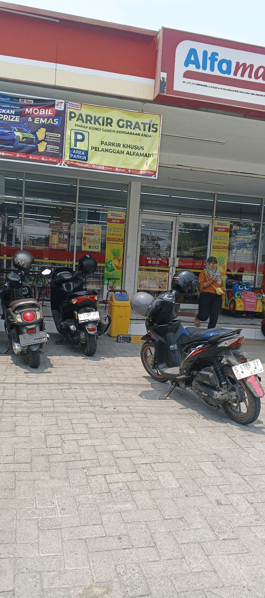 This is an outdoor, eye-level, full shot of an Alfamart convenience store in Sumberwudi, Indonesia. The facade of the store is red and white, with a prominent blue and white Alfamart logo. A large yellow banner in front of the store reads "PARKIR GRATIS" (Free Parking) and indicates that parking is for Alfamart customers.
In the foreground, parked on a brick-patterned sidewalk, are three scooters. The scooter on the left is silver with a red taillight. The middle scooter is black with a red taillight and a visible license plate "S 3187 JGD". The scooter on the right is black with a red rim on its rear wheel and a visible license plate "S 6779 JZ". A yellow trash bin is situated behind the middle scooter.
A woman wearing an orange t-shirt and dark pants is walking out of the store, holding a small yellow bag. Behind her, to the right, are colorful children's amusement rides. The store's entrance consists of glass doors, and through them, shelves with products are visible. The sky is clear and bright, suggesting a sunny day.