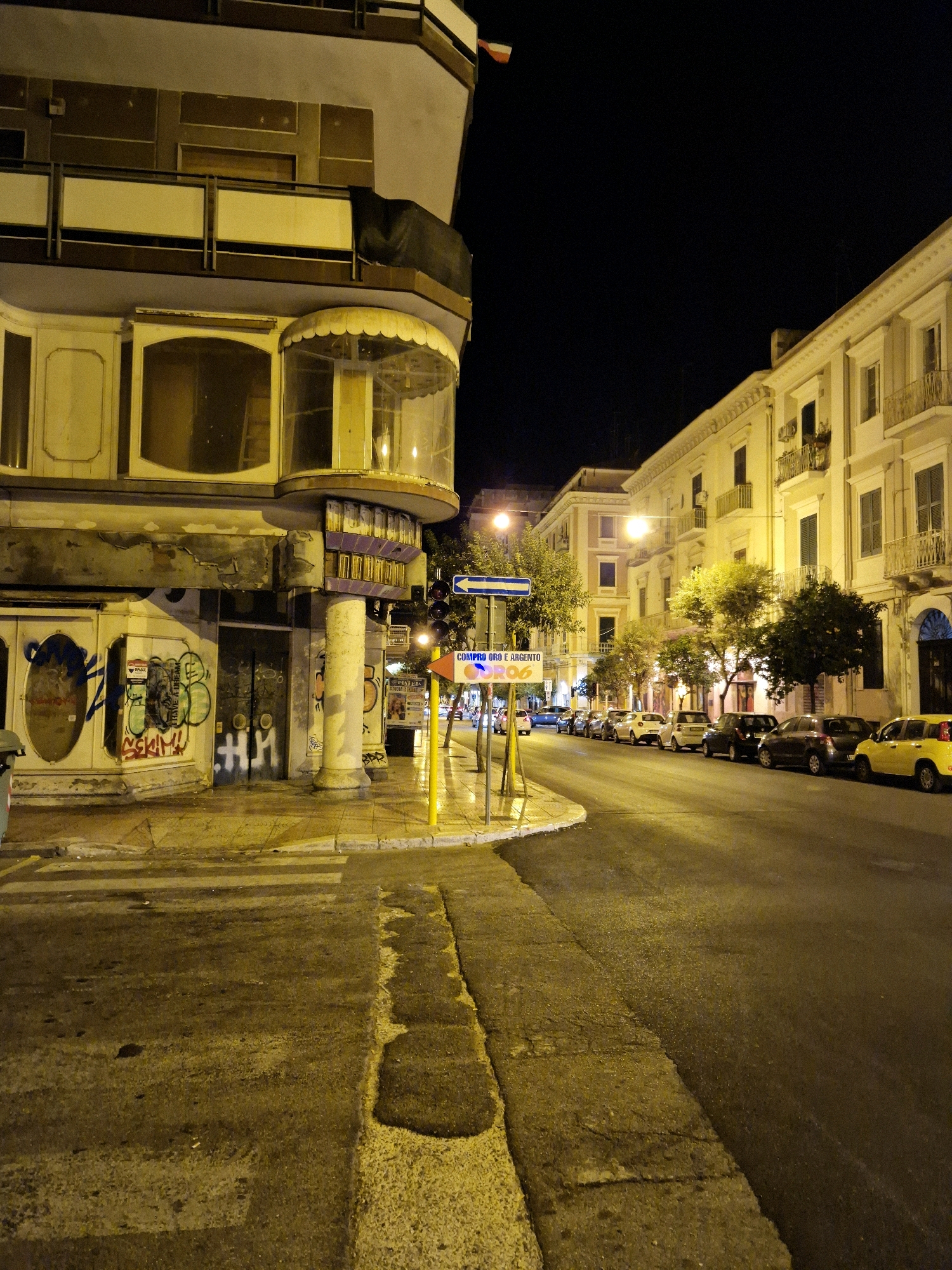 The photograph captures a nighttime street scene in Taranto, Italy, specifically on Via Giuseppe Verdi, near the intersection with Via Giuseppe Argento. The perspective is from street level, looking down the road, which is illuminated by streetlights, casting a warm, yellow glow on the wet asphalt and the surrounding buildings.
On the left side of the image, a building with a modern, curved facade dominates the foreground. Its lower level is covered in graffiti, including tags and what appears to be a stylized figure. Above this, there are balconies with railings and glass-enclosed sections. The building's architecture suggests it might be a commercial establishment or residential units with storefronts below.
The street itself is lined with parked cars, including a prominent yellow car on the right side. The road surface is dark and appears wet, reflecting the lights. The sidewalk on the right is paved with stone.
Across the street, other buildings are visible, characterized by their light-colored facades, balconies, and shuttered windows. These buildings appear to be multi-story residential or commercial properties.
A traffic light is visible on the right side of the street, along with directional signage indicating "COMPRO ORO E ARGENTO" (Gold and Silver Buying) and a blue directional arrow. A flag, appearing to be red and white, is affixed to a pole on the left side of the image, further up the street.
Trees line the sidewalk in some areas, their foliage illuminated by the streetlights, creating pockets of shadow and light. The overall atmosphere is one of a quiet urban night, with the yellow light creating a somewhat moody or vintage feel. No people are actively depicted in the scene. The image is a static, observational record of this urban environment at night.