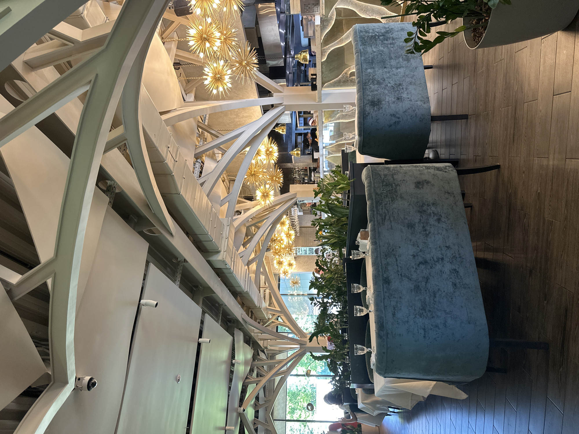 This image depicts the interior of an upscale restaurant or cafe, distinguished by its elegant and elaborate design. The dominant features are the ornate white architectural elements, including arches and lattice-like structures, that form a decorative ceiling or partition. Numerous spherical chandeliers, composed of many small light bulbs, hang throughout, casting a warm glow.
Seating consists of plush, teal-grey velvet upholstered banquettes, accompanied by tables set with crisp white tablecloths and clear stemmed glasses, suggesting readiness for dining. Potted green plants serve as decorative dividers between seating areas, adding a natural touch. The flooring is made of dark wooden planks laid in a tile pattern.
Through large windows, natural light and lush greenery are visible, indicating a daytime setting, though interior lights are also active. Several blurry figures of people, likely patrons or staff, can be seen in the background and near the windows. In the deeper background, behind a counter, another individual appears to be working.
Notable details include security cameras mounted on the walls. Visible text, seen in reverse through one of the windows, includes "CAXAP" and "CAXAPO," which are Russian words possibly indicating the establishment's name or a related theme (like "sugar"). The overall ambiance is sophisticated and inviting, designed for dining or social gatherings. The image is oriented sideways.