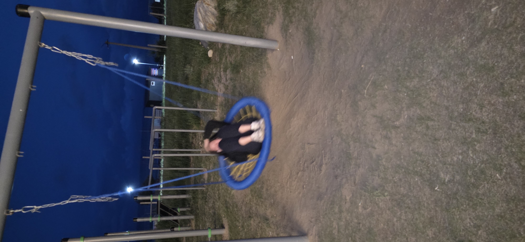 The image captures a young person on a circular swing at a playground in Bactu-Dolonotu, Kyrgyzstan, during twilight. The person, wearing dark clothing and white sneakers, is curled up on the blue and yellow woven swing, which is suspended by chains from a grey metal frame. The background features a few playground structures, including metal bars and poles, and beyond that, a hint of buildings and a single lamppost illuminated against the darkening blue sky. The ground is a mixture of dirt and sparse grass, suggesting a dry outdoor environment. The image's motion blur indicates the swing is in motion.