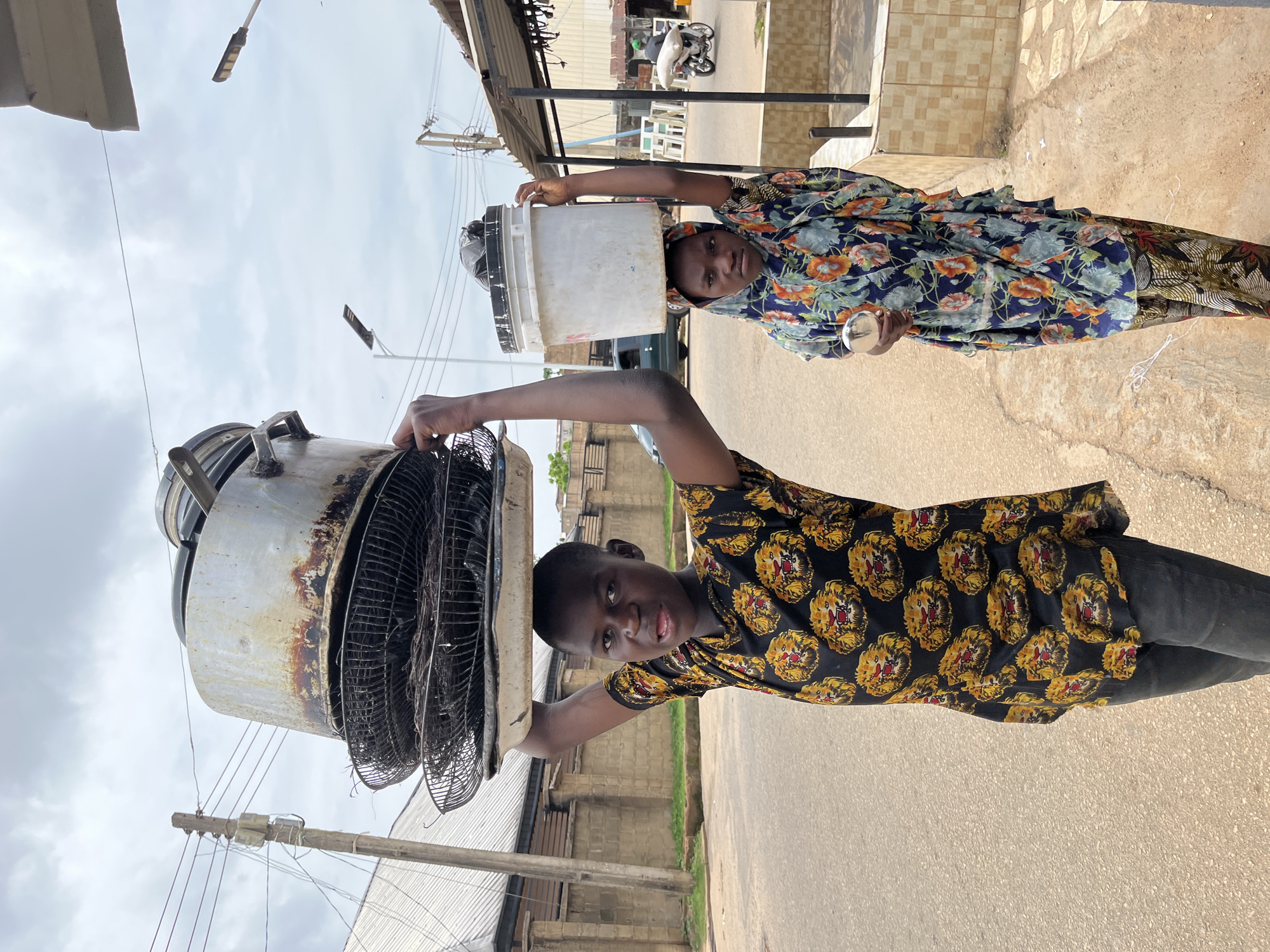 Two children are walking on a paved street in Jos, Nigeria, each carrying a large load on their head. The child in the foreground, a boy wearing a black t-shirt with a repeated pattern of lions, balances a stack of metal pots and wire baskets on his head. Behind him, a girl wearing a floral headscarf and dress carries a large white plastic bucket. The street is lined with buildings and utility poles, and the sky is overcast. A motorcycle is visible in the background. The overall scene depicts daily life in a busy urban environment.