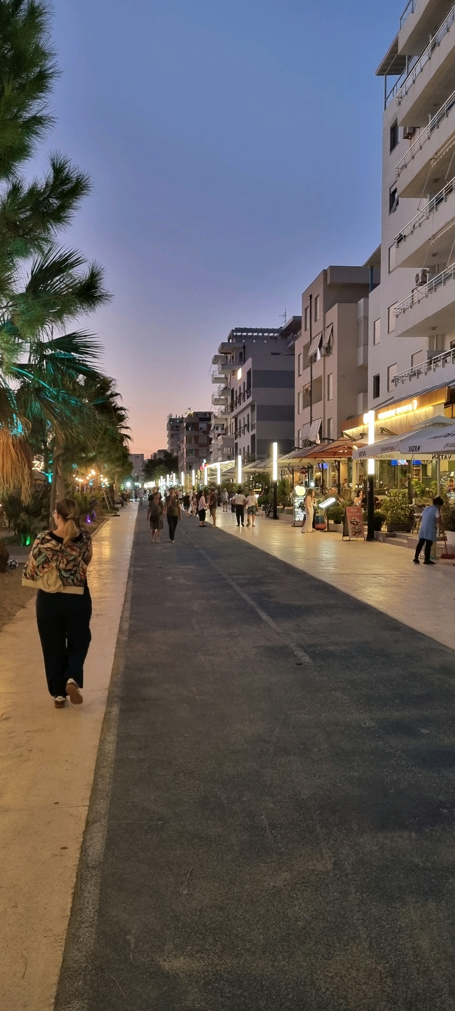 A woman with blonde hair tied back, wearing a floral print jacket and dark pants, walks away from the camera on a light-colored paved walkway. To her right is a dark asphalt road. Palm trees with decorative lights are visible on the left side of the walkway. Buildings with balconies line the right side of the road, with awnings and outdoor seating for restaurants and cafes. Several people are walking along the road and sidewalk. The sky is a gradient of deep blue to soft orange, suggesting it is dusk. The scene depicts a lively promenade in Bashkia Durrës, Albania.