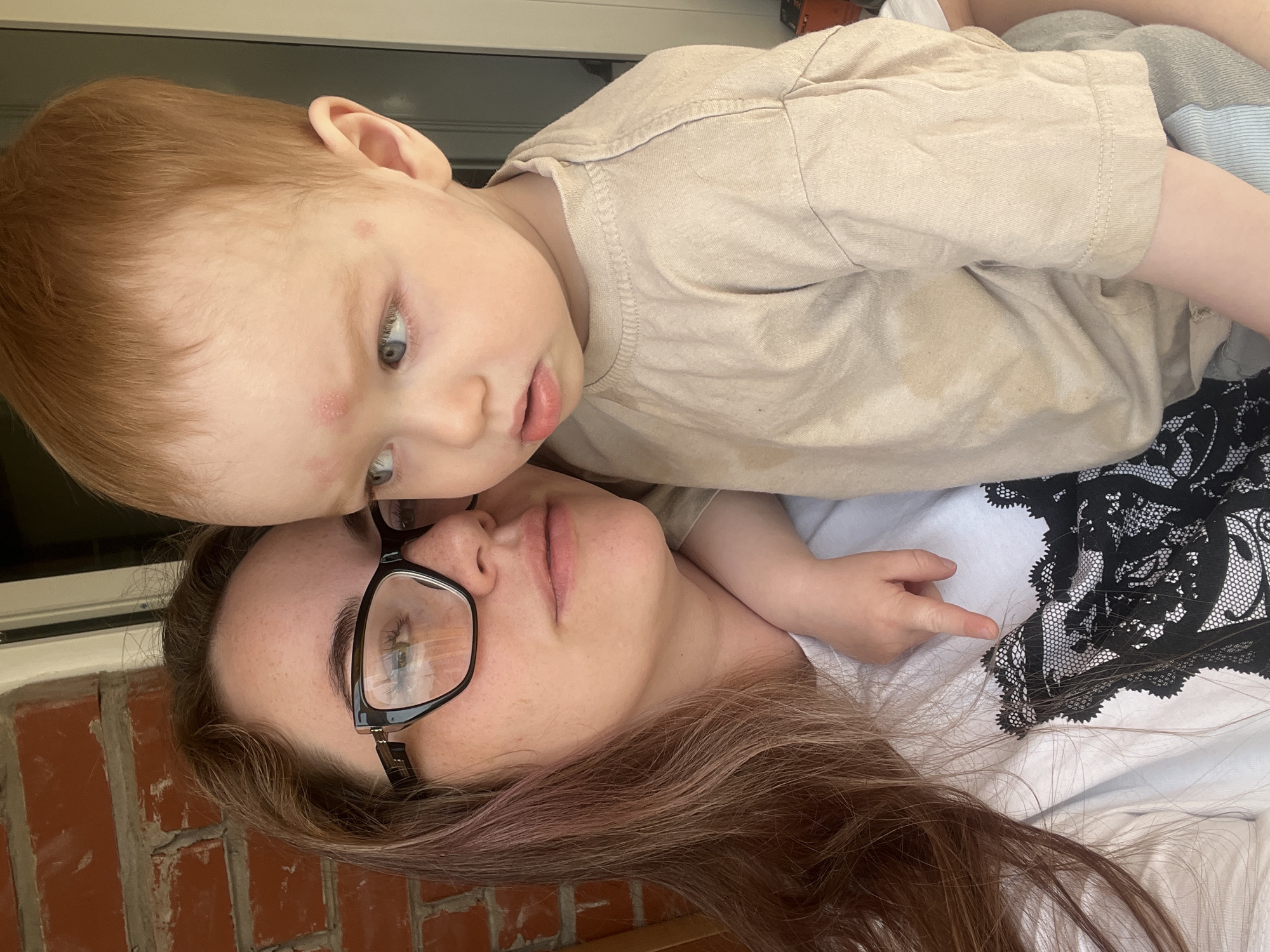 A young child with red hair and fair skin lies on top of a woman with long brown hair and glasses. The child is wearing a tan t-shirt. The woman is wearing a white t-shirt with a black lace design. They appear to be lying on a porch, as there is a brick wall and window in the background. The exact location in the city of Chaikovsky, Russia, cannot be determined from the image.