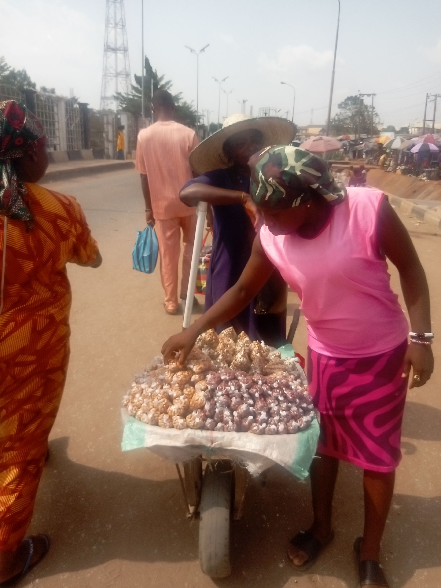 The scene depicts an outdoor urban environment in Onitsha, Nigeria, likely a street adjacent to a market. In the immediate foreground, a person wearing an orange and yellow patterned garment and a multicolored headscarf is partially visible from the back on the left. To their right, a single-wheeled cart or wheelbarrow, constructed with a wooden frame and a black tire, is laden with two distinct piles of small, roundish food items: one group is light brown/tan, and the other has a purplish tint, both resting on a light green fabric cover. A female individual, wearing a pink sleeveless top, a pink and magenta striped skirt, and a camouflage-patterned headscarf, stands next to the cart, leaning forward with her right hand extended towards the items. She wears a gold-colored ring on her right ring finger and multiple bracelets on her left wrist. Behind her, partially obscured, another person wearing a large straw hat and a dark garment is visible, with their arm resting on a white pole, possibly part of the cart. Further back on a dirt/sandy road, a male individual in a light orange shirt and trousers walks away from the camera, holding a light blue plastic bag in his left hand. In the background, a tall metal lattice tower is prominent against a hazy sky. Several streetlights are visible. To the left, a metal fence borders a property. To the right, further down the road, numerous market stalls with various colored umbrellas (red, white, purple) indicate an active market area.