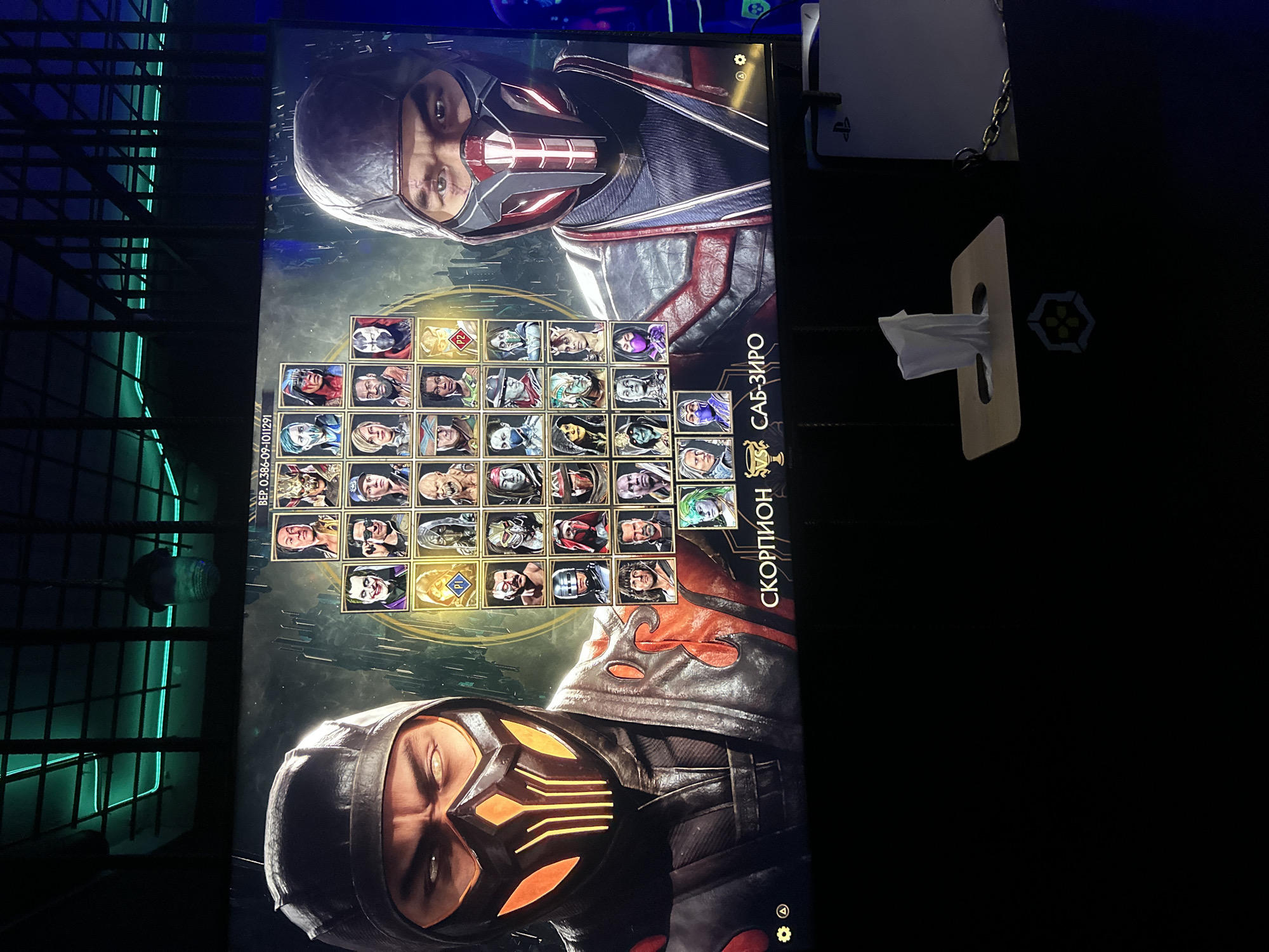 The image shows a screen displaying the character selection menu of the video game Mortal Kombat 11. The characters Scorpion and Sub-Zero are prominently featured on opposite sides of the screen. The game is being played on a PlayStation 5, which is visible next to the screen. A tissue dispenser is also nearby. The location is Yerevan, Armenia.