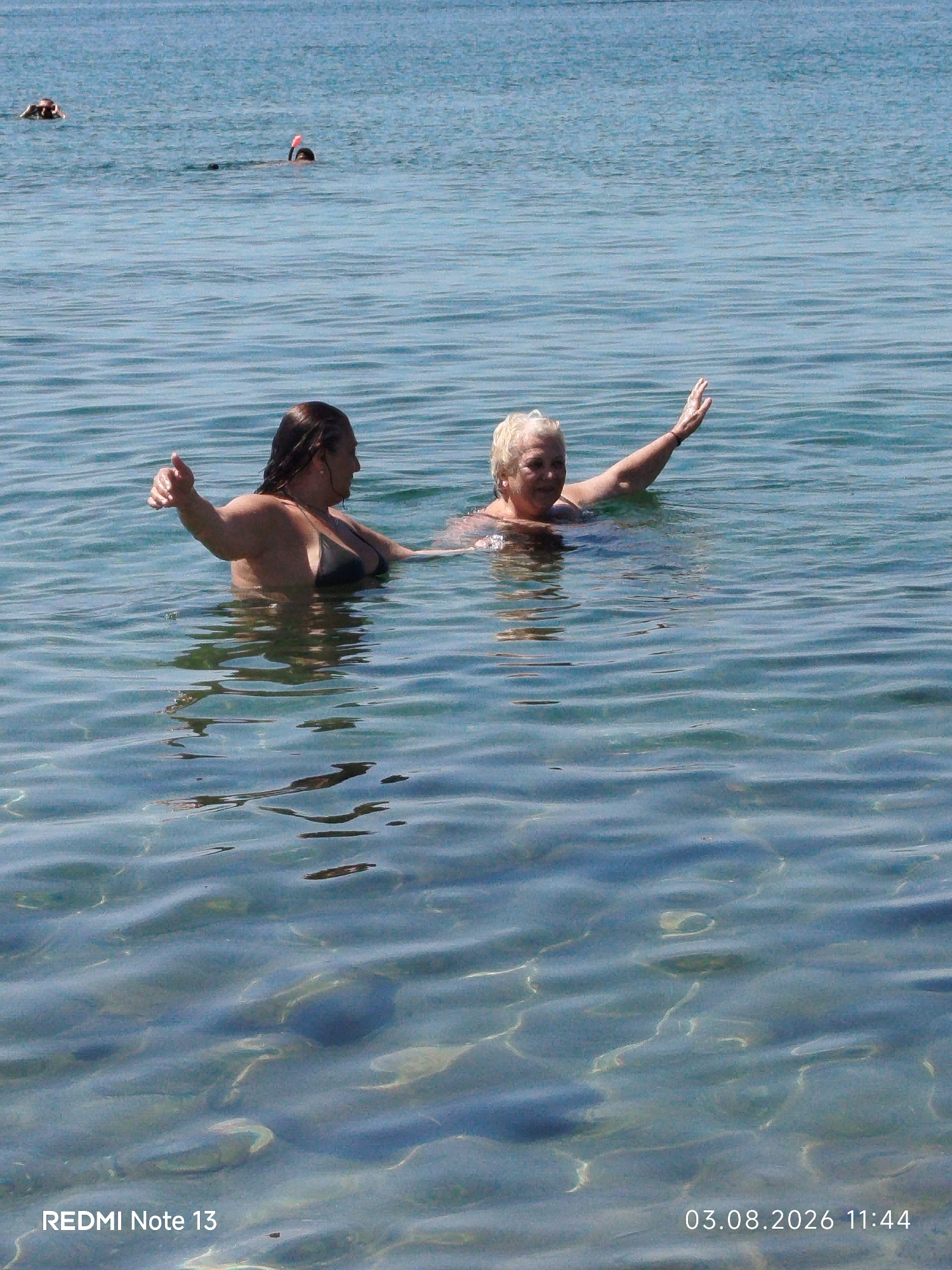 The image captures two women swimming in the clear blue water of Sithonia Municipal Unit, Greece. The woman on the left, with dark hair, is facing the right and appears to be giving a thumbs-up. She is wearing a dark bikini top. The woman on the right, with blonde hair, is facing the camera with her right arm raised, waving. She is also wearing a dark swimsuit. In the background, two individuals are visible in the distance, one with a snorkel mask. The water is shallow enough to see the sandy bottom with visible stones and light reflections. The timestamp "03.08.2026 11:44" and the text "REDMI Note 13" are superimposed at the bottom of the image.