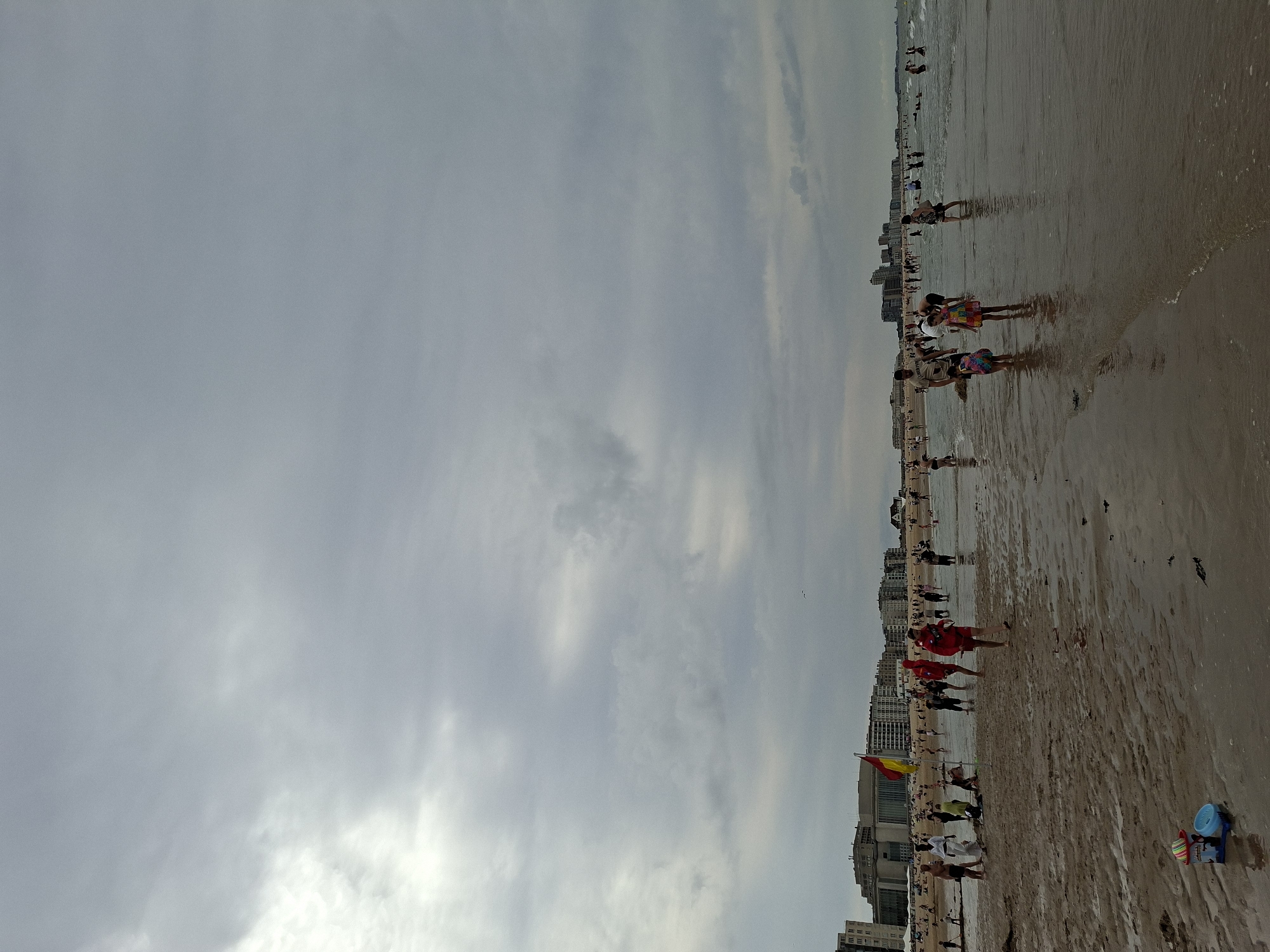 The image depicts a crowded beach scene in Ostend, Belgium, under an overcast sky. The foreground shows a wet sand beach with shallow water lapping at the shore. Several children's toys, including a bucket and spade, are visible in the lower left corner.
In the mid-ground, a large number of people are spread across the beach and in the shallow water. Some are walking, others are standing or playing. There is a distinct group of individuals wearing red, some of whom appear to be lifeguards, indicated by a red and yellow flag prominently displayed near them. Further back, more people are visible on the sand and in the sea, with some swimming or wading.
The background features a line of multi-story buildings, typical of a coastal city, suggesting an urban environment adjacent to the beach. The sky is heavily clouded, with patches of brighter light breaking through, creating a diffused lighting condition. The overall atmosphere is one of a busy summer day at the beach.