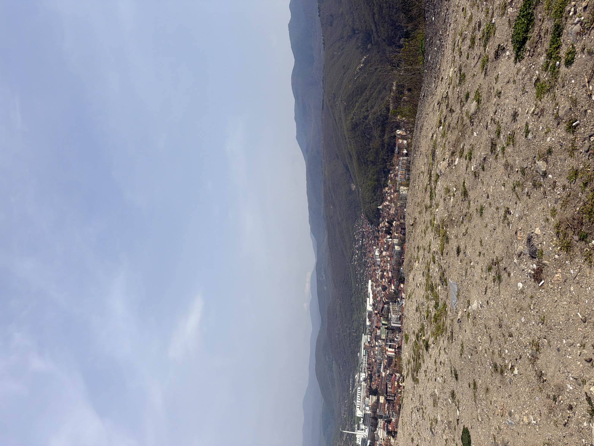 The image shows a landscape view in Sveti Konstantin, Bulgaria. The perspective is from an elevated, rocky, dirt area overlooking the town, which is nestled between a mountain range and what appears to be a slightly hazy sky. The town is densely built with a mix of low-rise buildings, and a few taller structures. The mountains are covered with dense vegetation. The ground in the foreground consists of dirt, rocks, and sparse patches of grass.