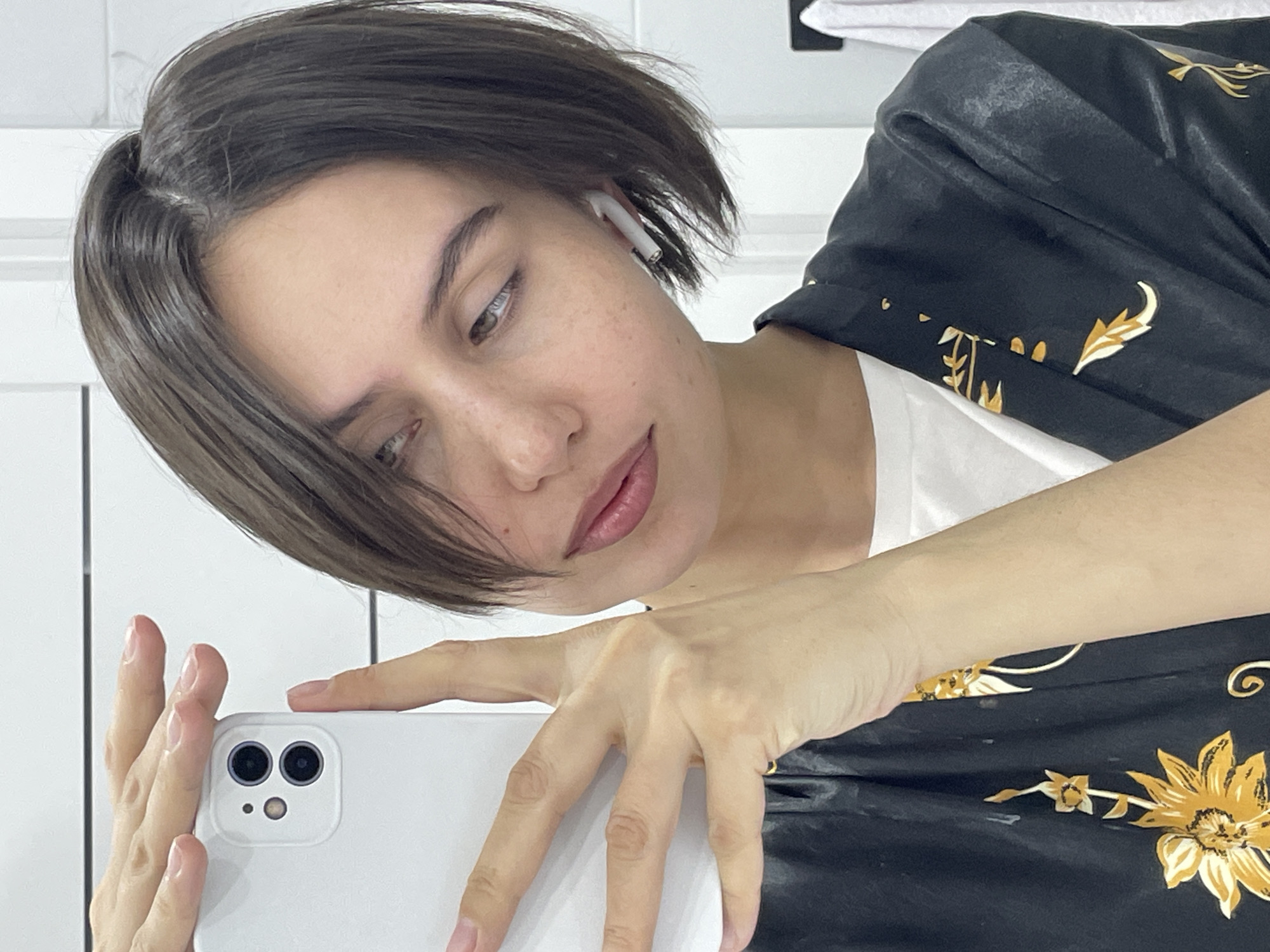 A female individual is depicted in a mirror selfie, framed from the chest up. She has short, dark brown hair, parted on the left side and swept across her forehead. Her skin is light-toned with visible freckles, and she possesses light-colored eyes and naturally pigmented lips. A white wireless earbud is present in her right ear. She is wearing a black robe or jacket adorned with gold and yellow floral and leaf patterns, over a plain white garment. Her left hand is prominently positioned, holding a white smartphone featuring two rear camera lenses and a flash, pointed towards the viewer. The background consists of white tiled walls with dark horizontal grout lines. A white towel hangs on a visible hook on the right side of the frame. No visual markers within the image indicate a specific location in Izhevsk, Russia.