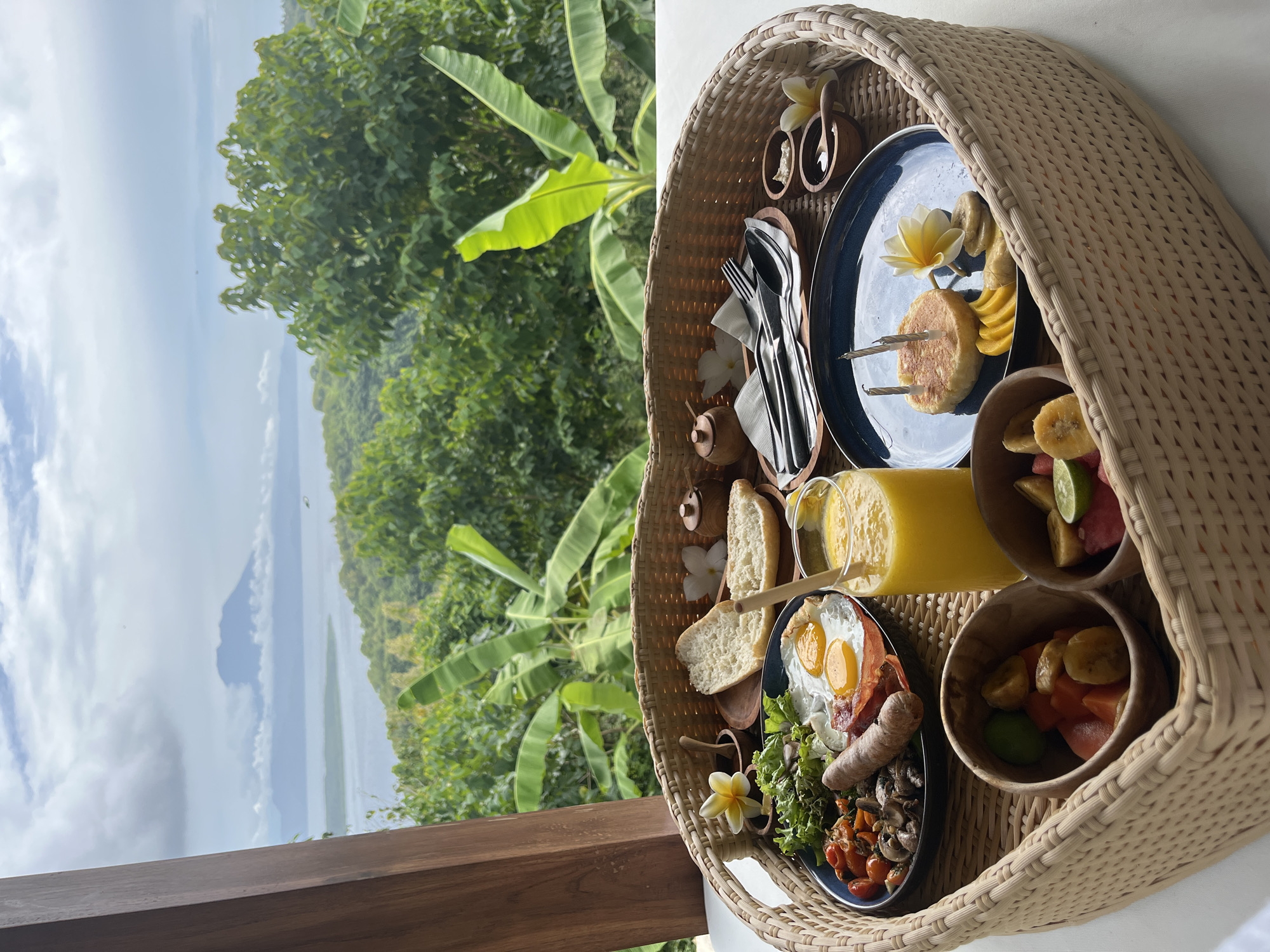 A lavish breakfast tray, laden with food, is captured in a photograph taken in Nusa Penida, Indonesia. The woven wicker tray holds an array of culinary delights: a plate with fried eggs, sausage, tomatoes, and mushrooms, along with another plate featuring what looks to be cakes or pancakes with fruit slices. A glass of orange juice and small wooden bowls filled with a mix of fruits, including bananas, papaya, and watermelon, complement the meal. Utensils, small condiments, and flowers add a touch of elegance to the presentation.
The setting is an outdoor balcony overlooking a lush green landscape, potentially a tropical forest or jungle, and a distant body of water, possibly the sea, under a cloudy sky. The wooden railing of the balcony is visible in the foreground, giving the perspective of a breakfast enjoyed with a scenic view.