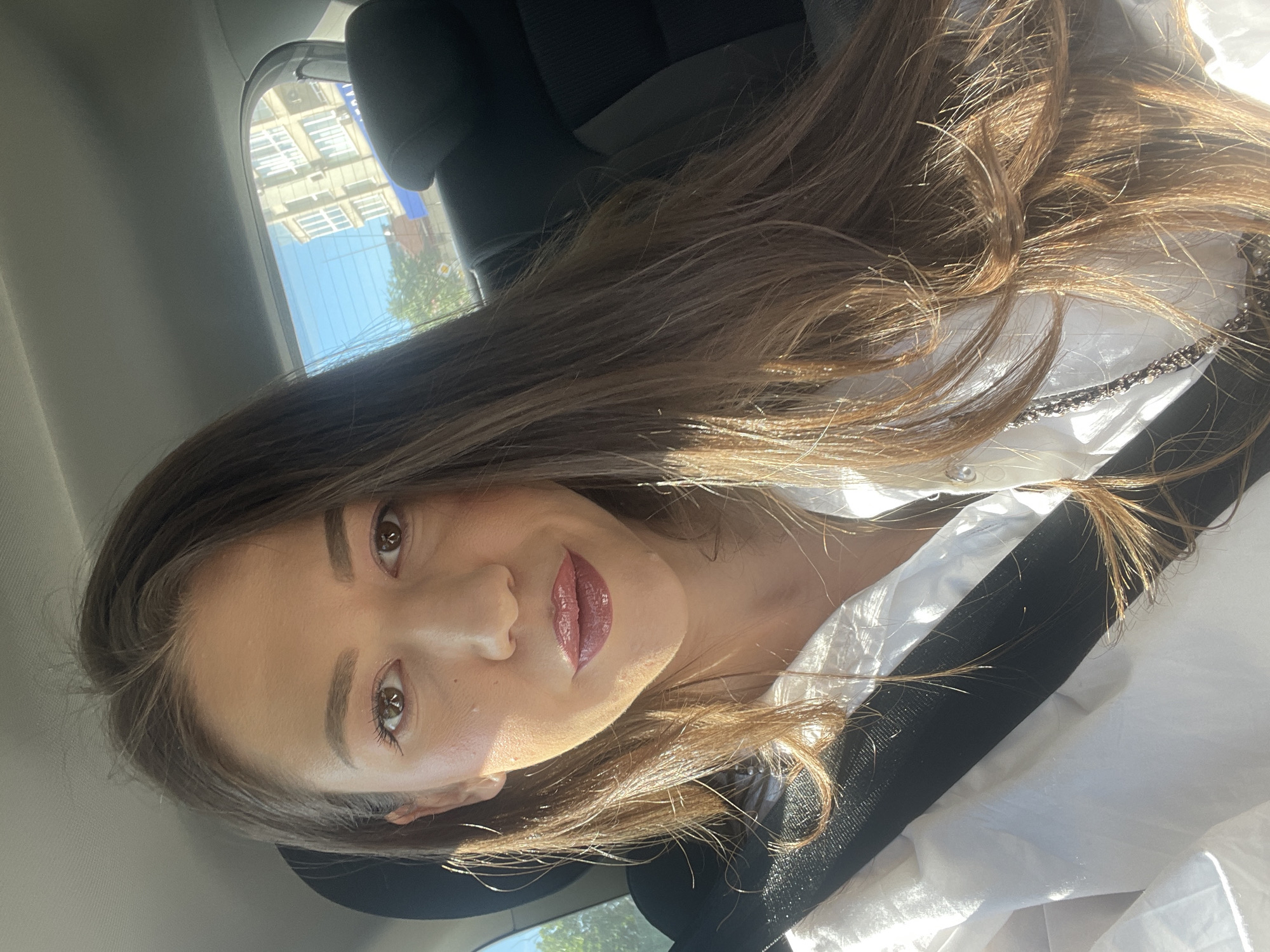 A woman is inside a car, facing the camera. She has brown hair, light skin, and is wearing a white shirt and a black seatbelt. Through the car window, a building with white window frames is visible, suggesting an urban setting. Based on the architectural style and context, the location is likely in the city of Sofia, Bulgaria. The woman appears to be in the passenger seat. The image captures a moment in transit, possibly during the daytime based on the natural light.