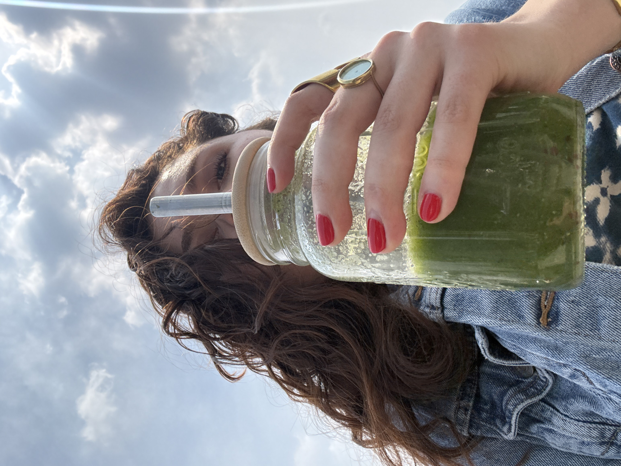 A low-angle shot captures a person holding a green drink against a bright, cloudy sky in Mexico City, Mexico. The individual, primarily seen from the forehead to the shoulders, has long, wavy brown hair and wears a blue denim jacket over a dark, patterned top. Their right hand, adorned with red nail polish and a distinctive gold ring featuring a circular light-colored stone, holds a textured glass bottle. The bottle contains a vibrant green liquid, likely a smoothie or juice, and has a light-colored lid from which a metal straw extends, positioned near the person's mouth area. The background is dominated by a clear blue sky with scattered white cumulus clouds, and a subtle lens flare indicates bright sunlight. The person appears to be drinking or about to drink from the bottle, enjoying a refreshing moment outdoors during the daytime. No visible text is present in the image.