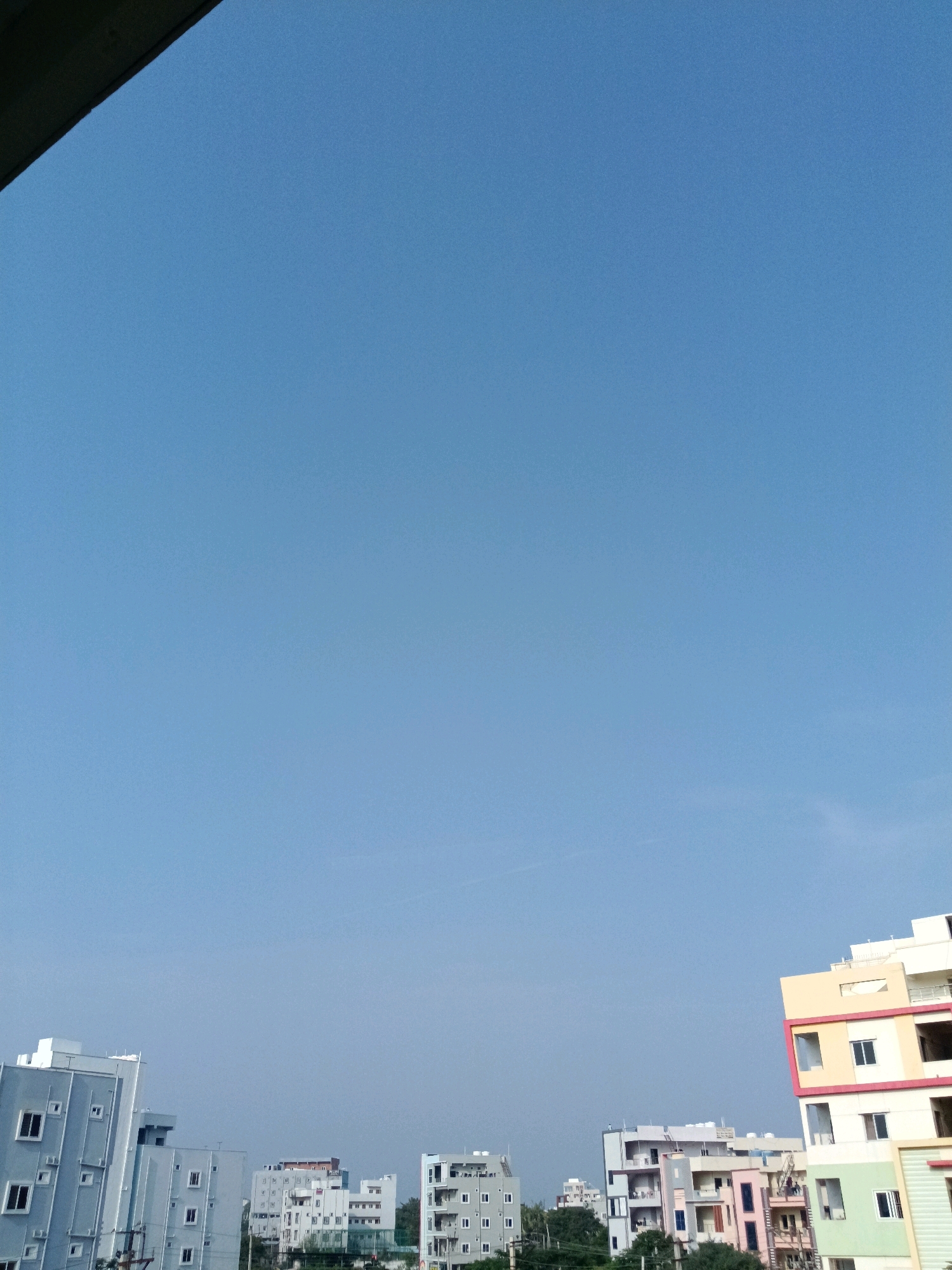 The image depicts a clear sky over a cityscape, likely Sheriguda, India, given the context. Several multi-story buildings are visible, characterized by their modern architecture and varying color palettes. In the foreground on the left, a light gray building with numerous square windows is prominent. To its right, another light gray building stands. Further into the background, a cluster of buildings displays a mix of white, pink, and light blue facades. On the right side of the image, a brightly colored building with yellow, red, and green accents is featured, its balconies and windows clearly defined. The sky is a uniform, bright blue, with a subtle haze visible in the distance, possibly indicating atmospheric conditions. The overall scene is one of an urban environment under a clear sky, with no visible people or specific activities taking place.