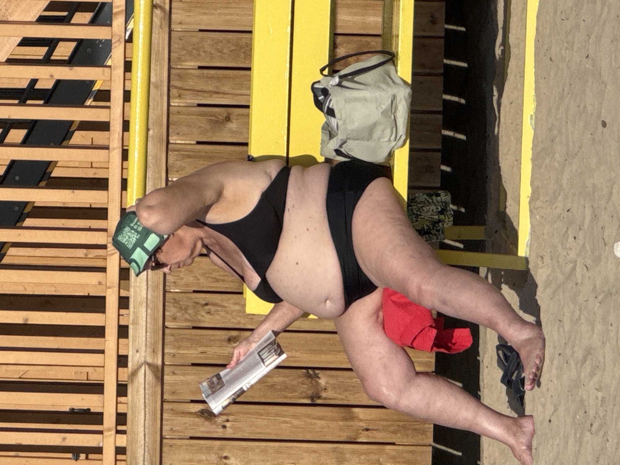 An elderly Caucasian woman wearing sunglasses and a green hat with a QR code pattern is sitting on a yellow bench on a sandy beach. She is wearing a black bikini and reading a magazine. A red towel is draped over her legs, and a light-colored bag is next to her on the bench. Black sandals are on the sand in front of her. The background consists of a wooden fence and wall. The location is a beach in Tallinn, Estonia.