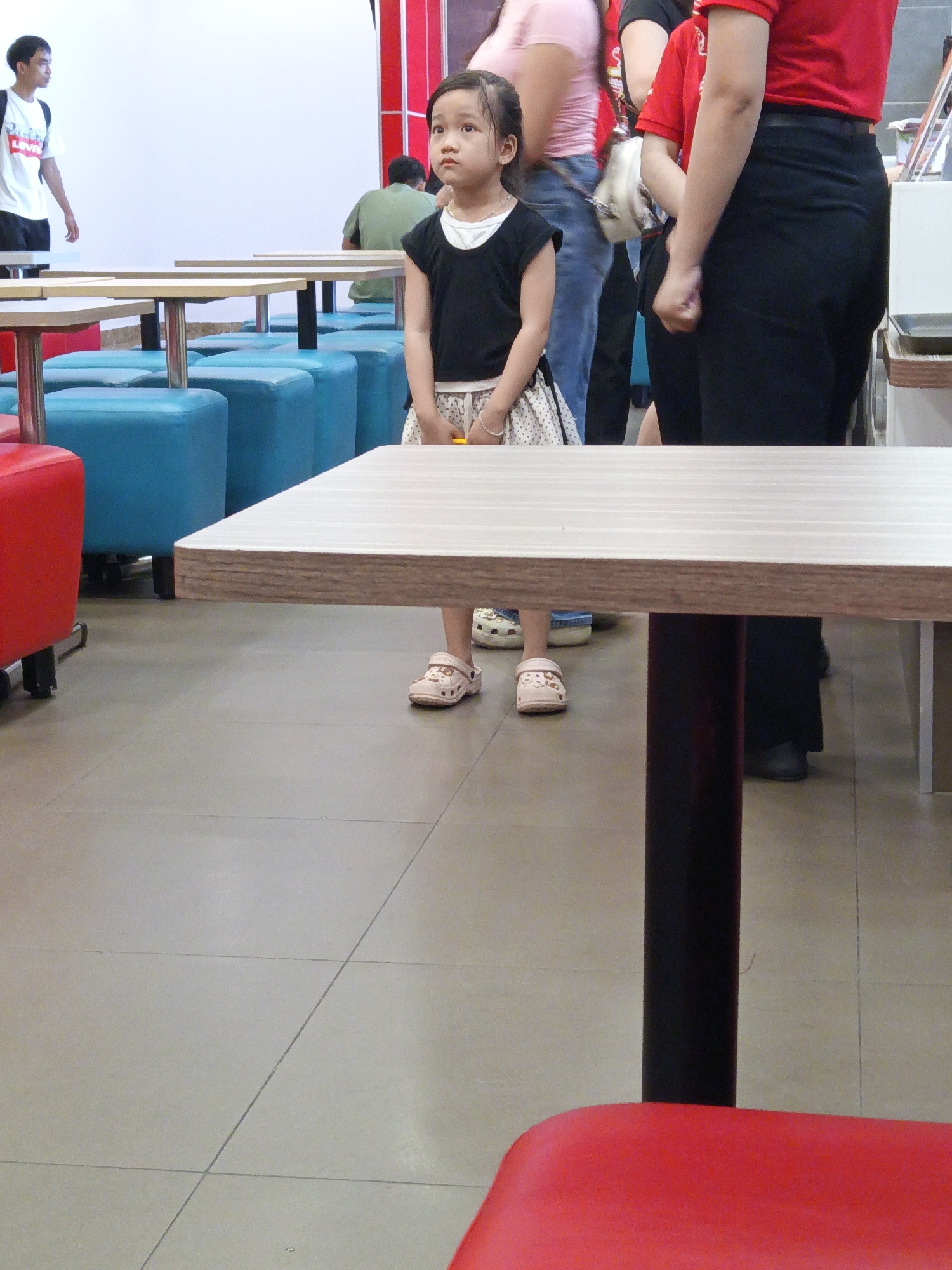 This media file depicts an indoor scene, likely a fast-food restaurant or a similar establishment, with a focus on a young girl standing in the foreground. The girl is wearing a black top and a patterned skirt, with light-colored sandals. Her gaze is directed upwards and to the left, suggesting she might be looking at someone or something outside the frame.
In the background, several other individuals are visible. To the left, a young man wearing a white t-shirt with red lettering and dark shorts is walking away from the camera. Further back, a person in a green shirt is seated. On the right side of the frame, an adult wearing a red shirt and black pants is partially visible, along with another person in a pink shirt. The setting includes multiple tables and blue seating benches. A red chair is prominent in the lower right corner of the image. The floor is tiled.
The lighting suggests it is daytime, and the indoor setting indicates protection from any potential weather conditions. The general atmosphere appears casual and public. There is no readily discernible text in the image, apart from the potential branding on the white t-shirt of the walking man, which is too blurry to read. The young girl's expression is neutral, and the overall impression is of a snapshot taken in a busy public space. The location context of Nha Trang, Vietnam, suggests this could be a dining area within that city.