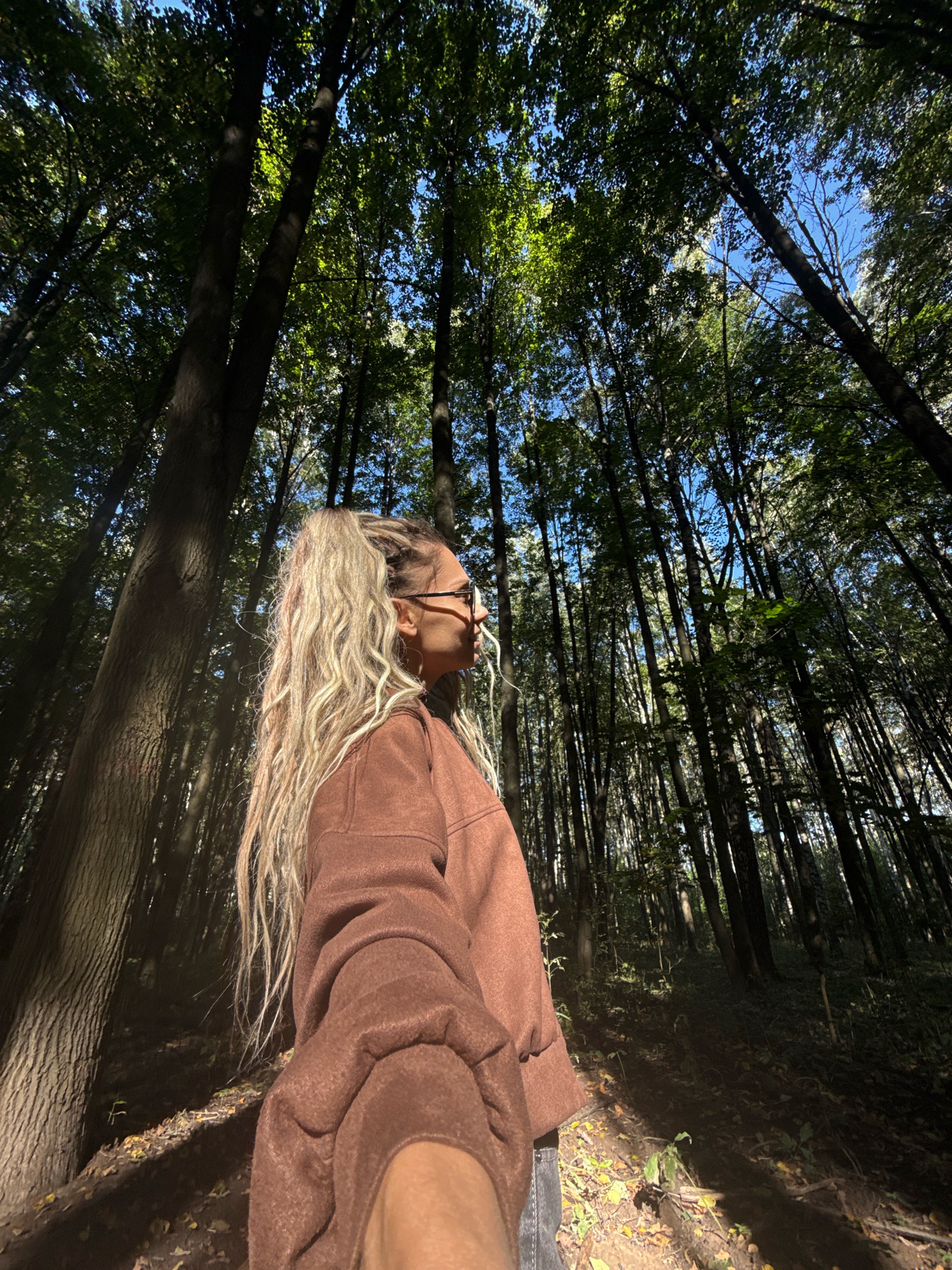 A low-angle outdoor photograph captures a young woman standing in a forest, facing to her right and looking up. She has long, blonde, wavy hair tied in a high ponytail and is wearing glasses and a brown fuzzy long-sleeved shirt. Her left arm is extended toward the camera. The background is dominated by tall trees with green leaves, creating a dense forest canopy with patches of blue sky visible through the foliage. The ground is covered with fallen leaves and dirt. The lighting suggests it is daytime, with sunlight filtering through the trees, casting shadows. The scene appears to be in a natural, wooded environment, consistent with a location in or near Rusanovo, Russia.
