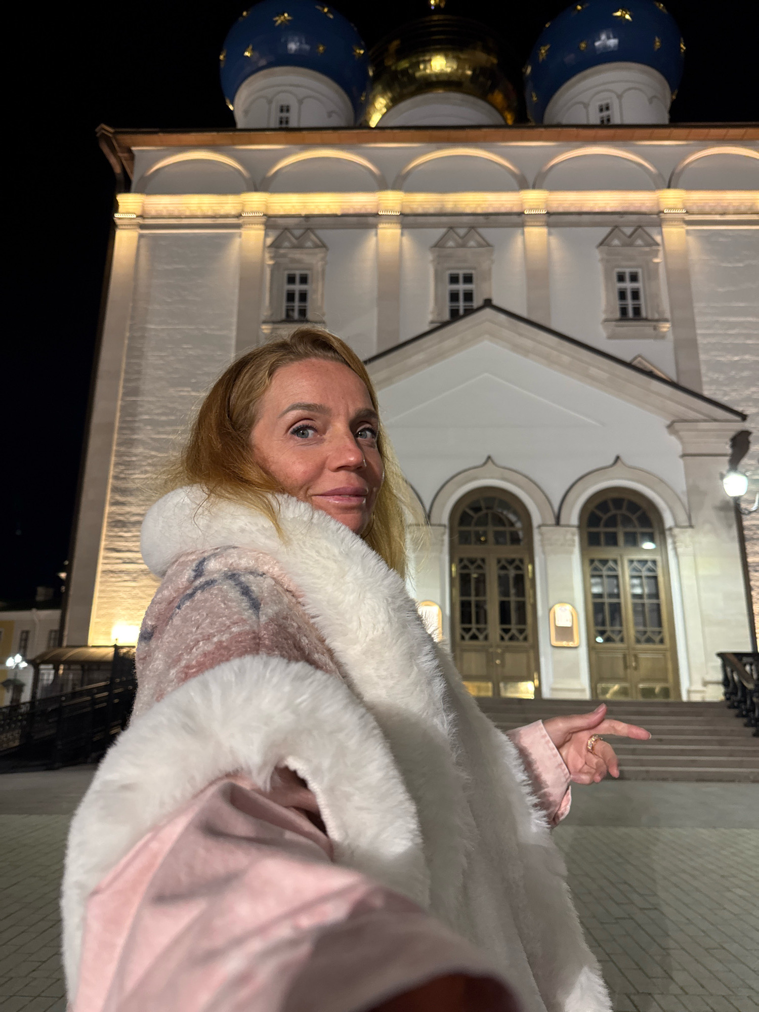 A woman with blonde hair is standing outside a building at night. The building is white with arched doorways and windows. There are blue domes with gold stars on top of the building. The woman is wearing a pink and white coat with a fur collar. She is looking at the camera and smiling. Her right hand is raised and her index finger is pointing. The ground in front of the building is paved with bricks. The scene appears to be in the city of Tver, Russia.