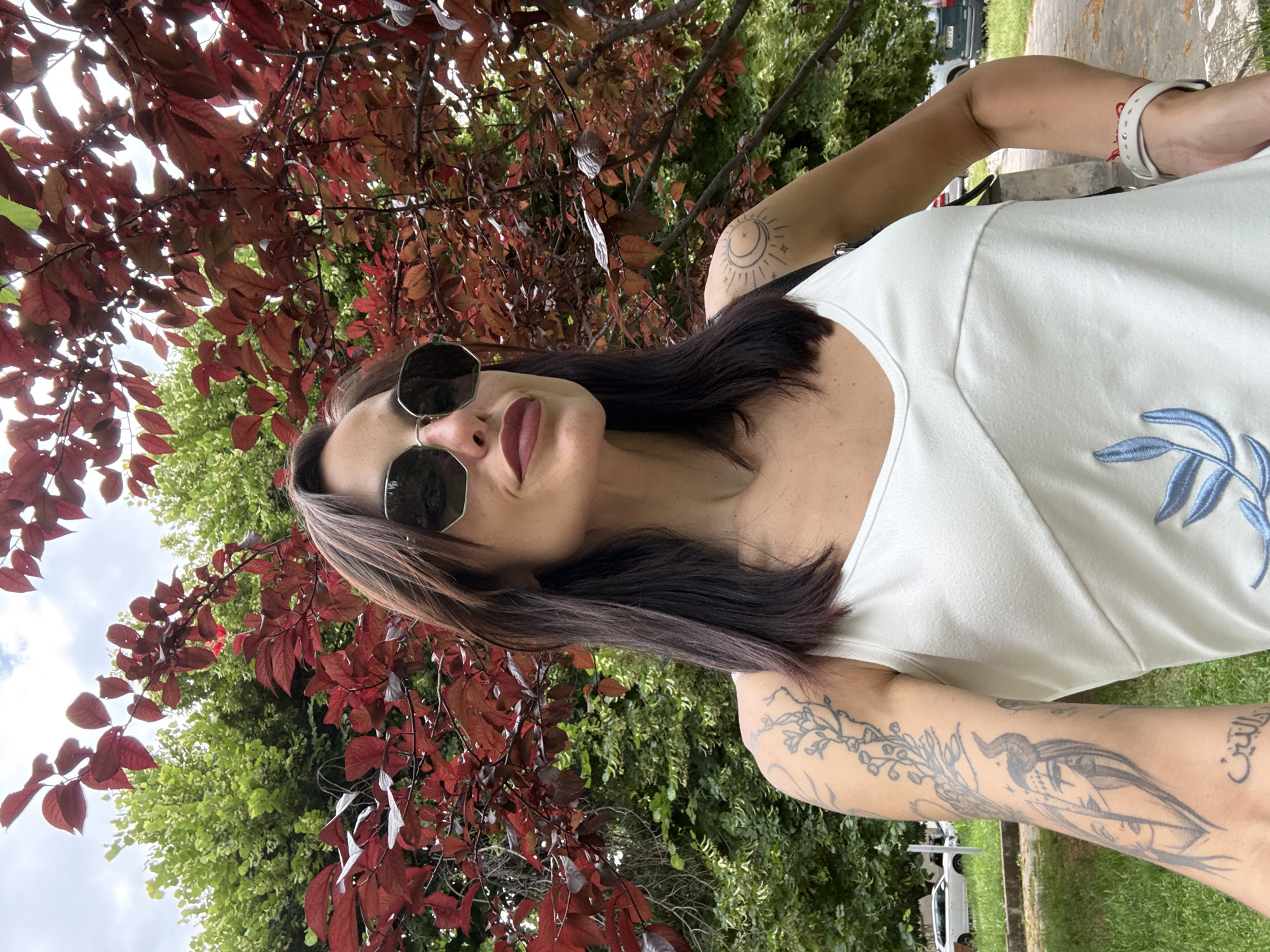 This is a close-up, waist-up, selfie-style photograph of a woman outdoors. The woman has dark hair, sunglasses, and is wearing a white sleeveless top with blue embroidery. She has several tattoos on her visible arm, including one of a moon and sun, and another of a woman with horns. She is standing in front of a tree with dark red leaves and a background of green trees and foliage. There are some cars parked in the background, on what appears to be a street or parking lot adjacent to a grassy area. The lighting suggests it is daytime, possibly overcast. The location appears to be a park or residential area within the city of Pleven, Bulgaria, given the presence of the cars and the general urban greenery.