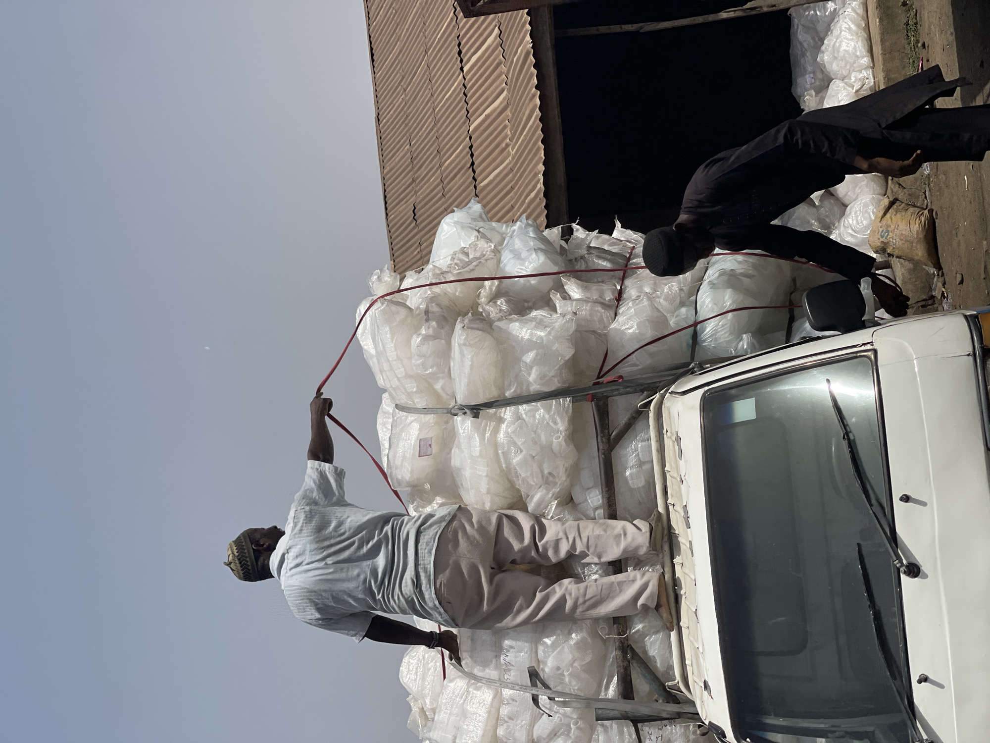 In Zaria, Nigeria, two men are atop a white truck loaded with tightly packed, white plastic bags. The bags appear to be stacked high, secured with red ropes. One man, wearing a light-colored shirt, tan pants, and a patterned cap, is adjusting a rope, while the other man, dressed in black, appears to be leaning over, possibly securing the load or unloading it. The truck is parked next to a building with a corrugated metal roof. The sky is overcast.