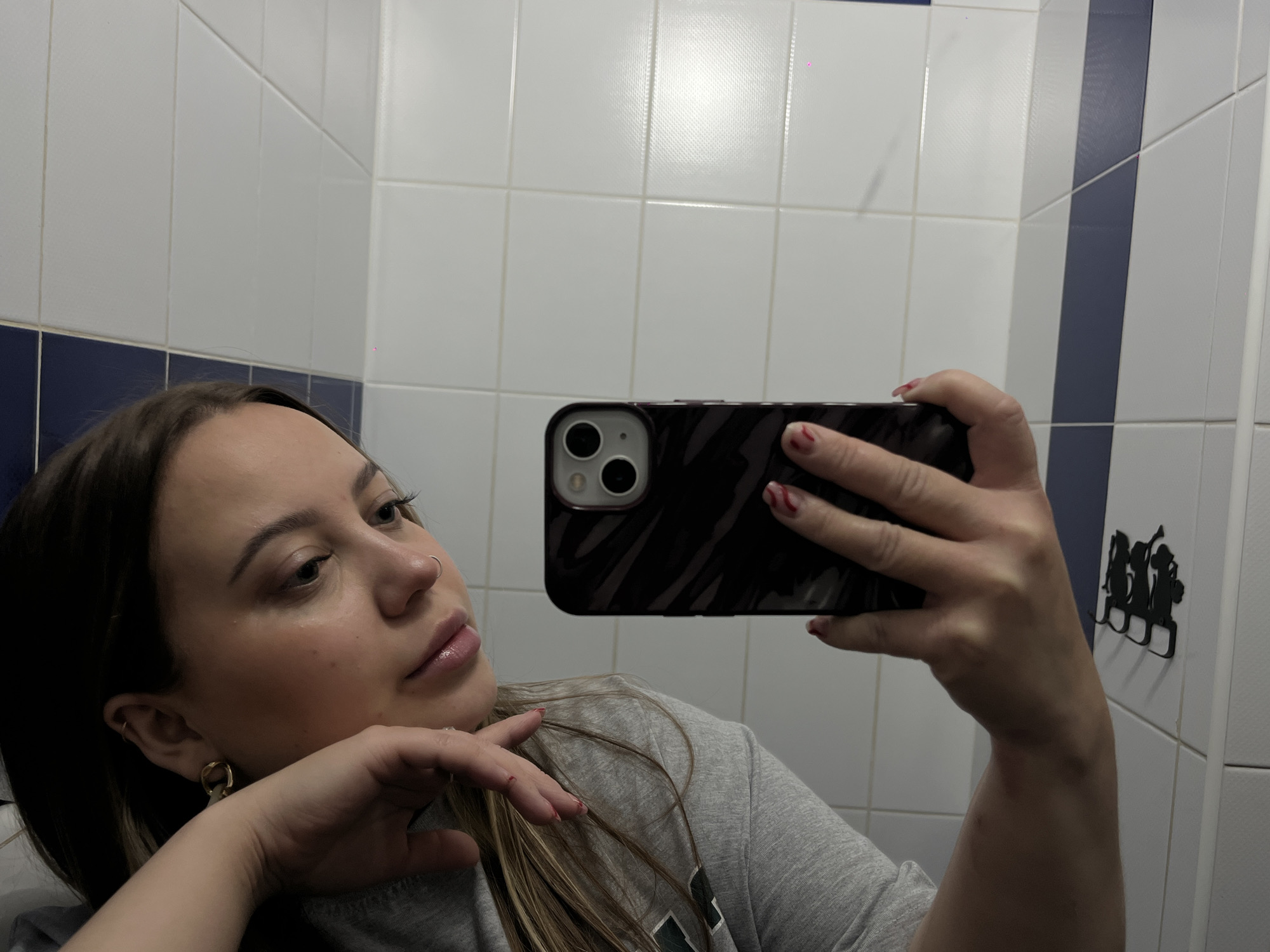The scene depicts a mirror selfie taken by a female individual in an indoor environment, specifically described as Novopokrovskaya, Russia. The subject, a woman with long, dark brown hair featuring lighter streaks, wears a grey short-sleeved t-shirt. Her right hand, adorned with red and white striped nail polish, holds an iPhone, partially obstructing her face. The phone has a dark, patterned case and prominently displays its two rear camera lenses. Her left hand, also with red and white striped nail polish that appears slightly smudged, rests against her jawline, supporting her head as she looks towards the phone/mirror. She wears a gold-colored hoop earring in her right earlobe and a smaller hoop in the upper helix, along with a silver-colored nose ring in her left nostril. Visible makeup includes blush/bronzer and lip gloss. The background consists of white ceramic tiles with light grey grout lines. A horizontal strip of dark blue tiles runs across the lower wall, and a vertical strip of dark blue tiles is visible on the right side of the frame. Artificial overhead lighting is reflected in the upper tiles. To the right of the subject, a black, wall-mounted hook rack, featuring three stylized female figures, is affixed to a white surface.