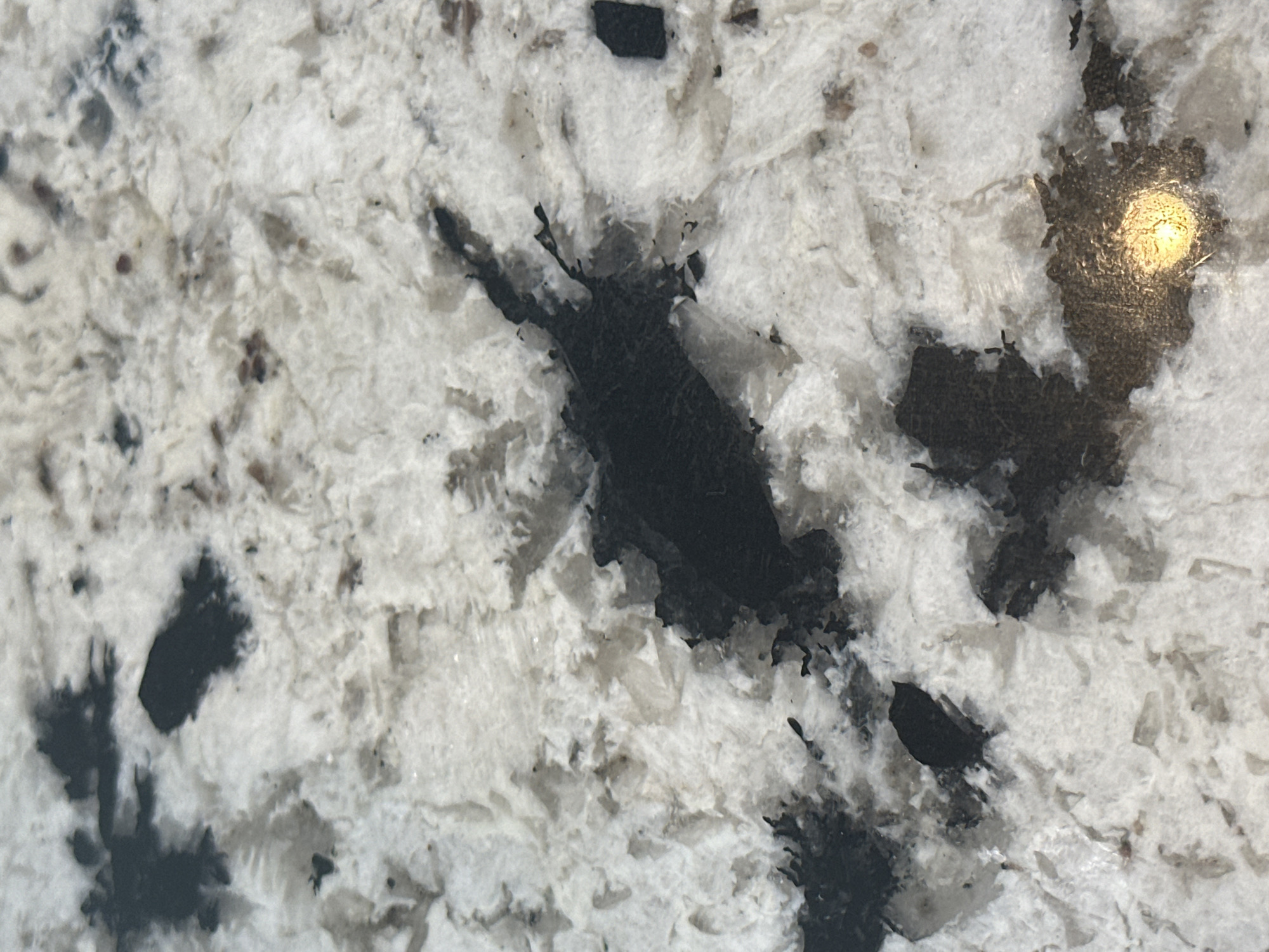 The image displays a close-up, overhead view of a speckled granite countertop, characterized by a pattern of black and white crystalline formations. The surface exhibits varying textures, with some areas appearing smooth and others rougher, suggesting a natural stone material. There is a distinct, dark, irregular shape in the center of the frame, possibly a vein or inclusion within the granite. A small, bright yellow-orange reflection, likely from a light source, is visible on the lower right portion of the surface, illuminating a section of the dark pattern. The overall impression is that of a durable, decorative surface.
This image does not contain sufficient information to determine its location in Burnaby, Canada, or any details about people, actions, or interactions.