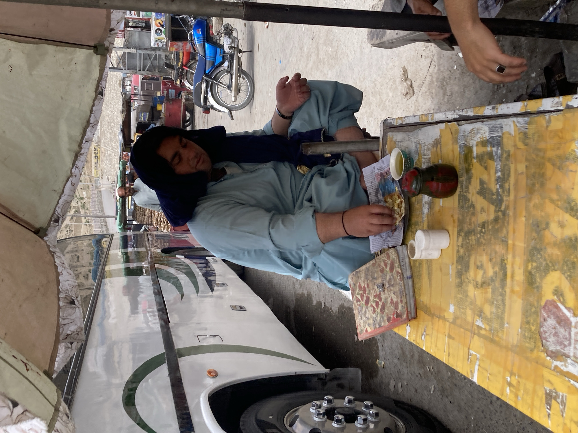 A man in a light blue shalwar kameez and a dark blue hooded garment is resting or sleeping, seated beside a weathered yellow table with peeling paint. His eyes are closed, conveying a sense of peace. On the table are a red thermos, a white cup, a piece of food resembling an omelet served on newspaper, a book with a red and white patterned cover, and two small white cylindrical objects.
The scene appears to be an outdoor area, likely a bus stop or roadside eatery in Quetta City Tehsil, Pakistan. To the left, part of a large white and green long-distance bus is visible, including its wheel. In the background, a blue Suzuki motorcycle is parked, along with a red vehicle. A beige canvas canopy provides shade over the area. Faintly visible in the distance are hills. Another person's hand, wearing a ring, is seen in the upper right, and the head of another individual is partially visible behind the resting man. The lighting suggests it is daytime with soft, indirect sunlight. Faint Urdu script and the partial English word "LOCK" can be seen on signs in the background. The man's attire and the general setting are consistent with the specified location.