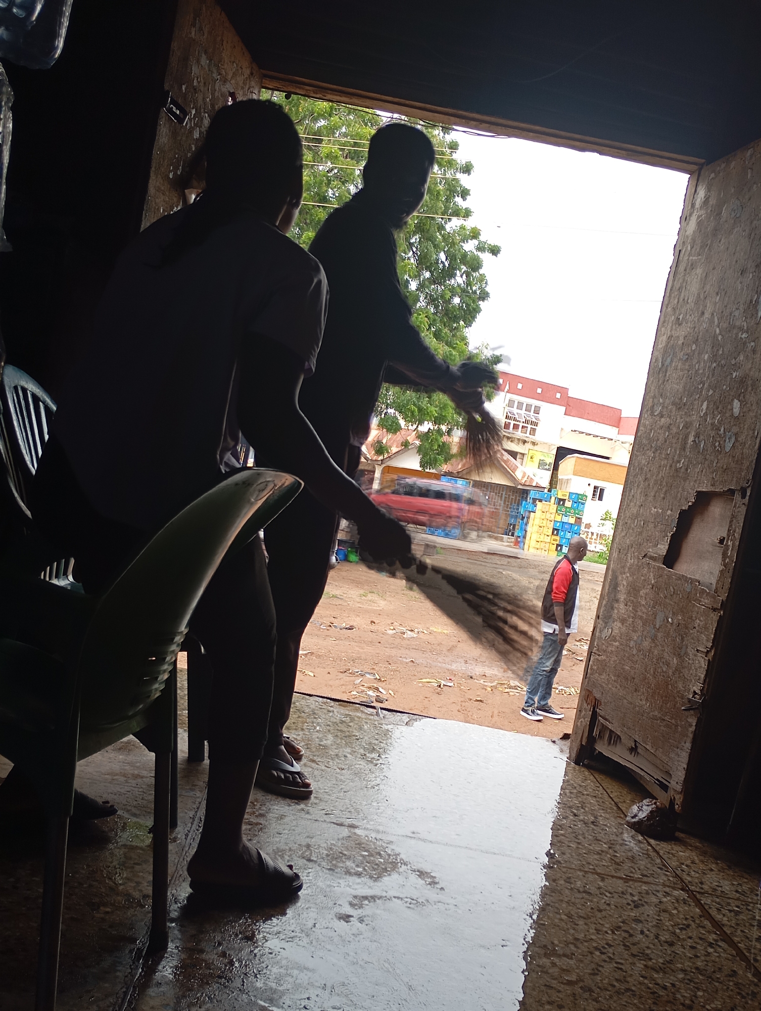 The image depicts an outdoor scene at what appears to be a shop or open-air market in Shendam, Nigeria. Two individuals are silhouetted in the foreground, one on the left holding a broom and actively sweeping, their body angled towards the right. Another silhouetted figure is to their right, also facing towards the right and appearing to observe the sweeping. In the middle ground, a third person, a male, stands facing away from the camera, looking towards a building with several stacks of colorful crates behind him. The ground is wet and reflective, indicating recent rain. The background shows buildings and trees, with a red truck visible to the left of the crates. The overall lighting suggests it is daytime, possibly overcast. The scene conveys a sense of daily activity in a commercial setting.
