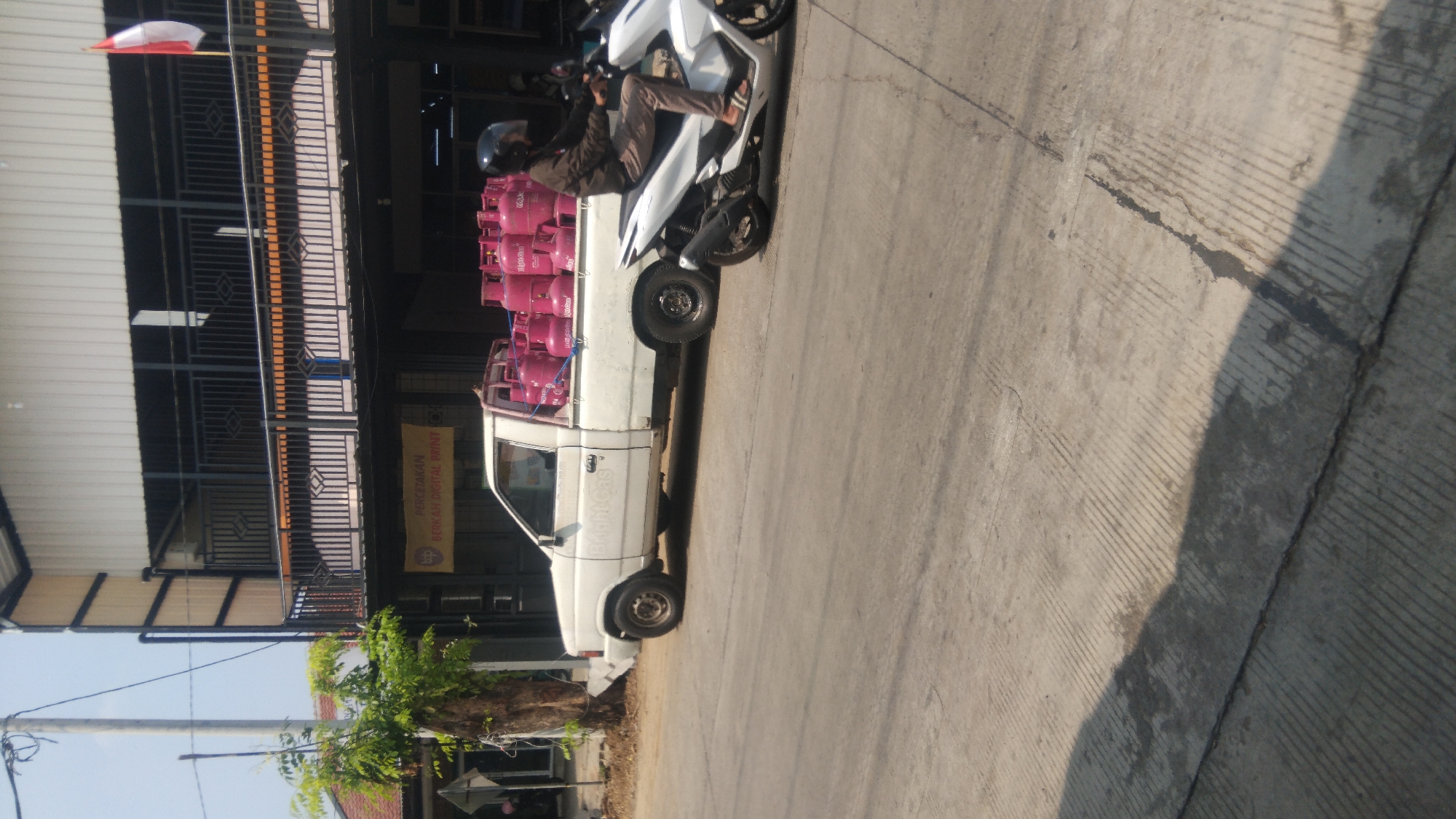A white pickup truck loaded with pink propane gas cylinders is parked on the right side of a street in Batang, Indonesia. A man wearing a brown jacket and helmet is riding a white motorcycle to the right of the truck. The truck has "Bright Gas" written on its side. The street is paved with concrete and has visible tire tracks. The background shows a two-story building with a balcony and a sign that reads "PERCETAKAN BERKAH DIGITAL PRINT". A flag of Indonesia is also visible on the building. The sky is clear and the sun is shining, casting shadows on the ground.