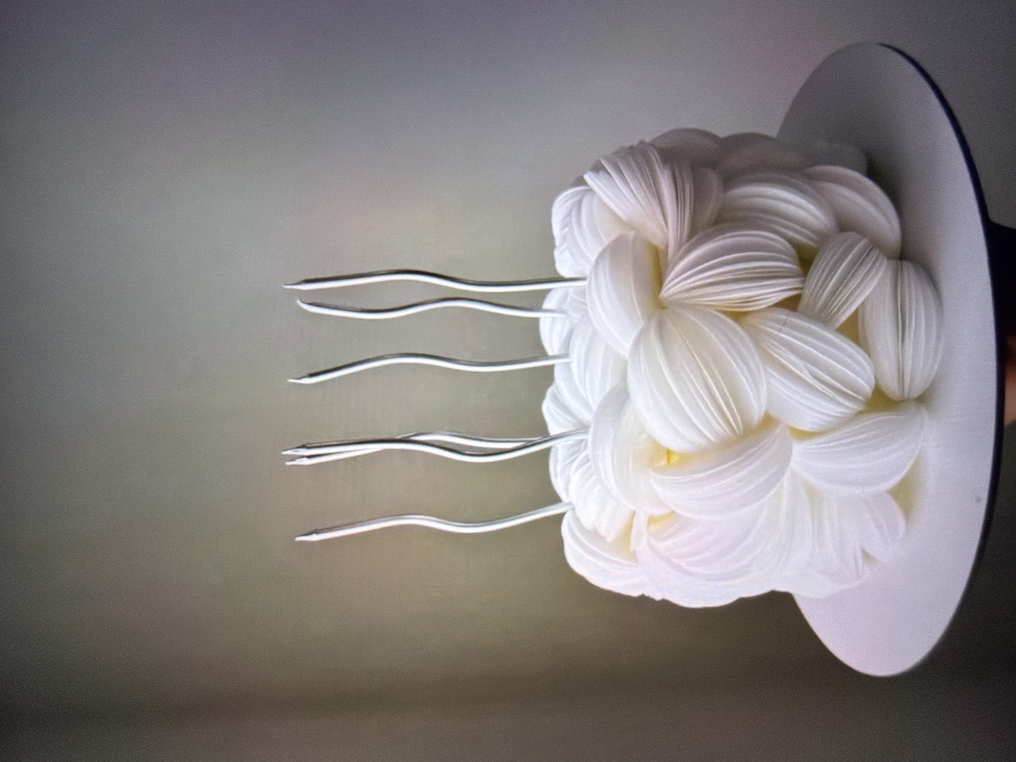 The image shows a close-up, eye-level shot of a small, white, elaborately decorated cake. The cake is cylindrical and covered in delicate, petal-like folds of frosting or fondant, giving it a textured, almost floral appearance. Emerging from the top of the cake are four thin, silver, wavy candle holders, empty. The cake sits on a plain white cake stand, which is itself on an unseen surface. The background is a gradient of soft, muted colors, transitioning from a darker, grayish-brown on the left to a lighter, purplish-gray on the right, suggesting a dimly lit or neutral indoor environment. There are no people or animals visible in the image. The scene is devoid of any specific location markers that would tie it to Beșalma, Moldova.