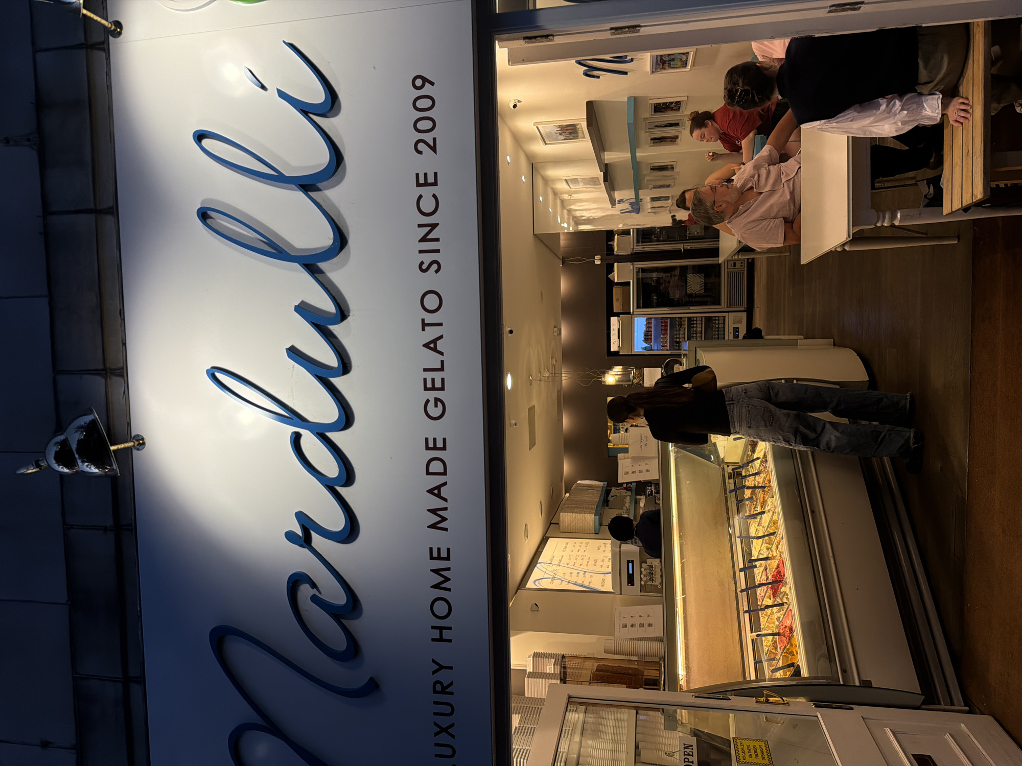 The image depicts the storefront of "Nardulli," a gelato shop in London, United Kingdom. The name and the phrase "LUXURY HOME MADE GELATO SINCE 2009" are displayed on the front. Inside the shop, several customers are seated at tables, while another person is standing at the gelato display case, presumably making a purchase. The gelato case shows a variety of flavors. A sign near the open door reads "OPEN".