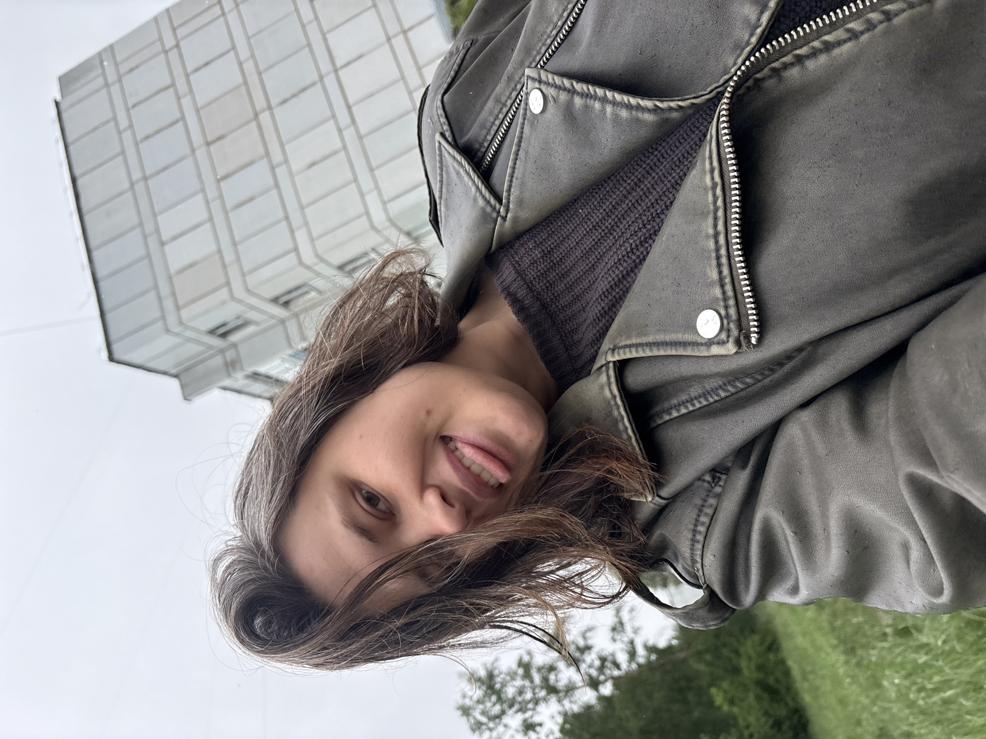 A woman is looking into the camera, sticking out her tongue. She is wearing a leather jacket and a dark sweater. Behind her is a building and some trees. The location is Chaikovsky, Russia.