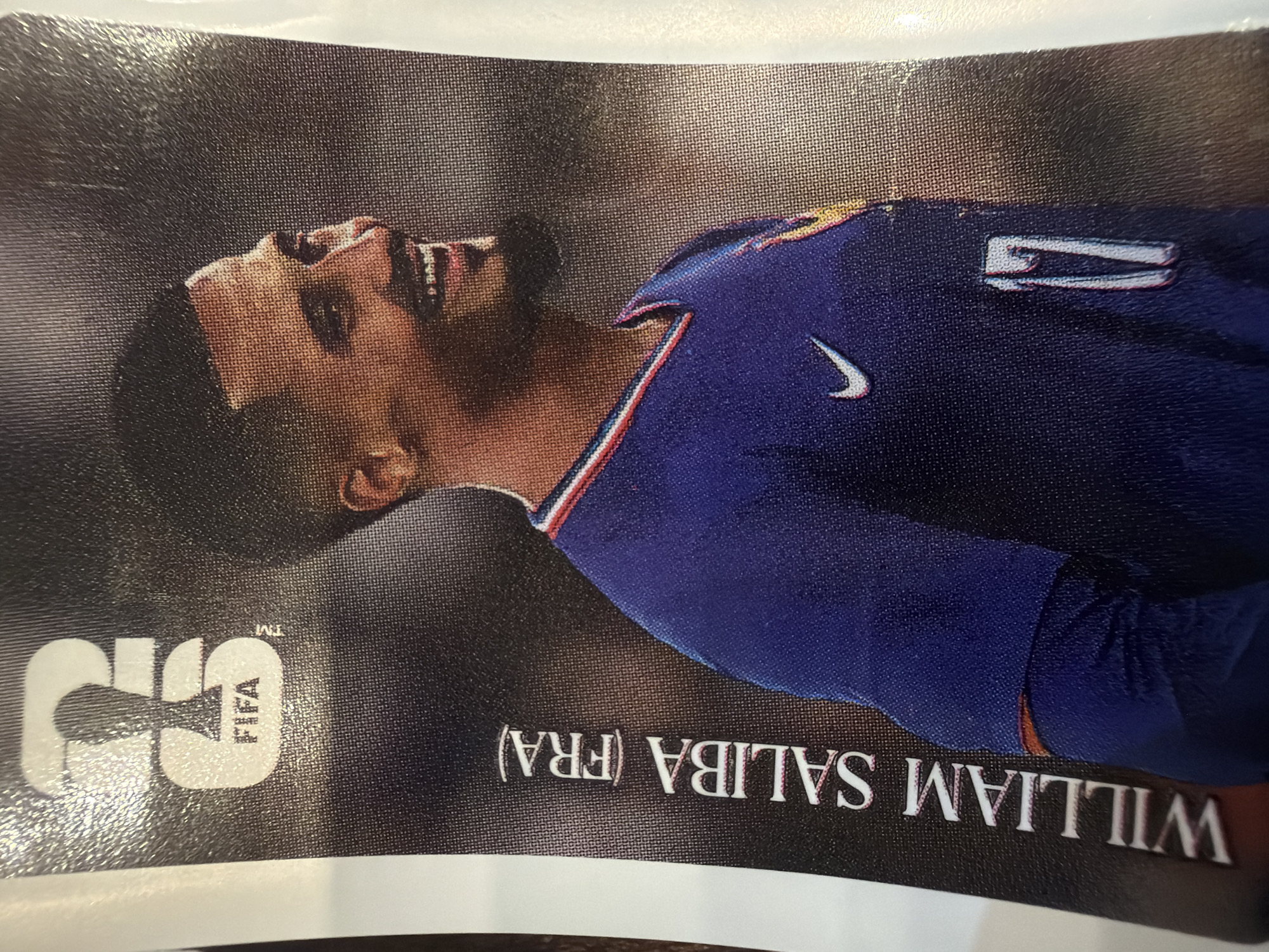 A close-up, vertical, full-body shot shows a young adult male soccer player named William Saliba from France, wearing a blue jersey with white Nike logo, the number 17, and red and white trim around the neck. The player has short, dark hair and is smiling. The FIFA logo is visible in the upper left corner. The image is a trading card or sticker.