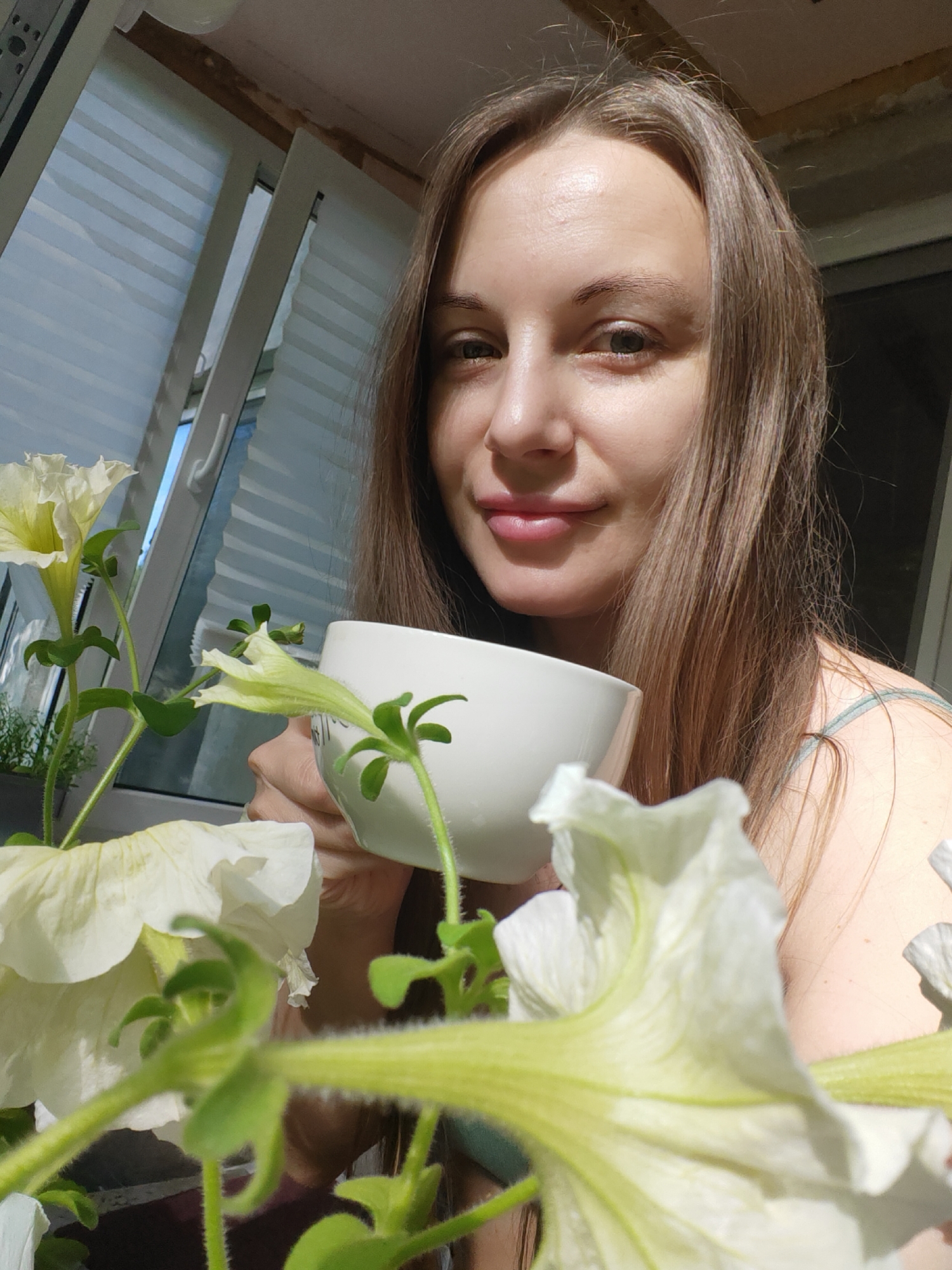 A woman is holding a white mug with some flowers in the foreground, possibly a self-portrait or a photo taken by someone close. The woman has light brown hair and is looking directly at the camera with a slight smile. The mug is partially obscuring her face, and the flowers are arranged around the mug, creating a natural frame. The background shows a window with white blinds, suggesting an indoor setting. The photo was possibly taken in the city of Chaikovsky, Russia, based on the query context. The lighting appears to be natural, possibly from the window.