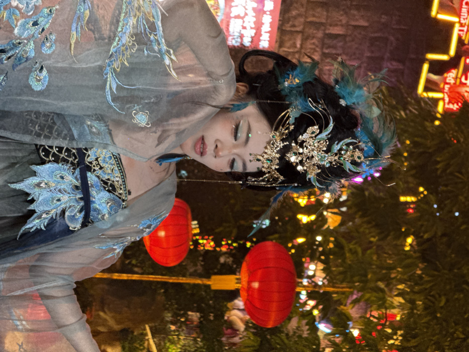 A woman in a traditional Chinese Hanfu dress and elaborate hairpiece stands in front of red lanterns and illuminated foliage. The dress is adorned with intricate embroidery in shades of blue, silver, and gray. The hairpiece is decorated with feathers and jewels. Her makeup includes sparkly eyeshadow and defined eyebrows. The background is blurred, with hints of buildings and streetlights.