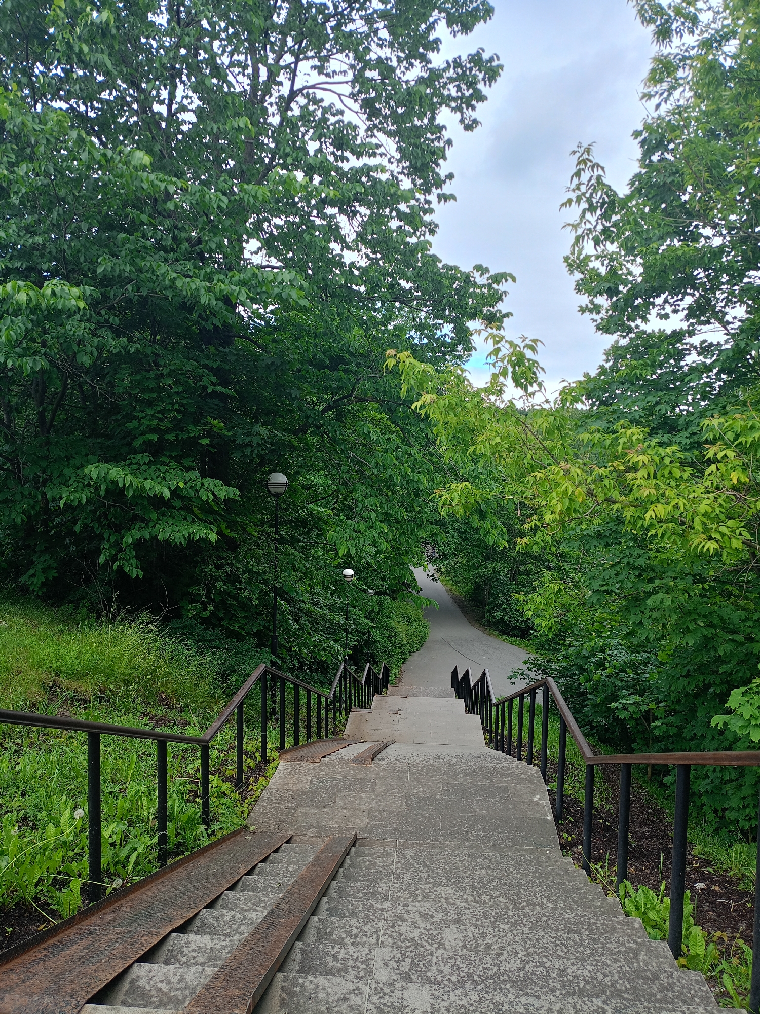 A wide, concrete staircase with metal handrails and ramps descends from the foreground into a tree-lined park. The stairs are bordered by lush green grass on the left and a path with more stairs and handrails on the right. The path is paved and winds through dense green foliage. Two lampposts are visible along the path. The sky is overcast with some visible clouds. The overall impression is one of a peaceful, natural setting within a city.