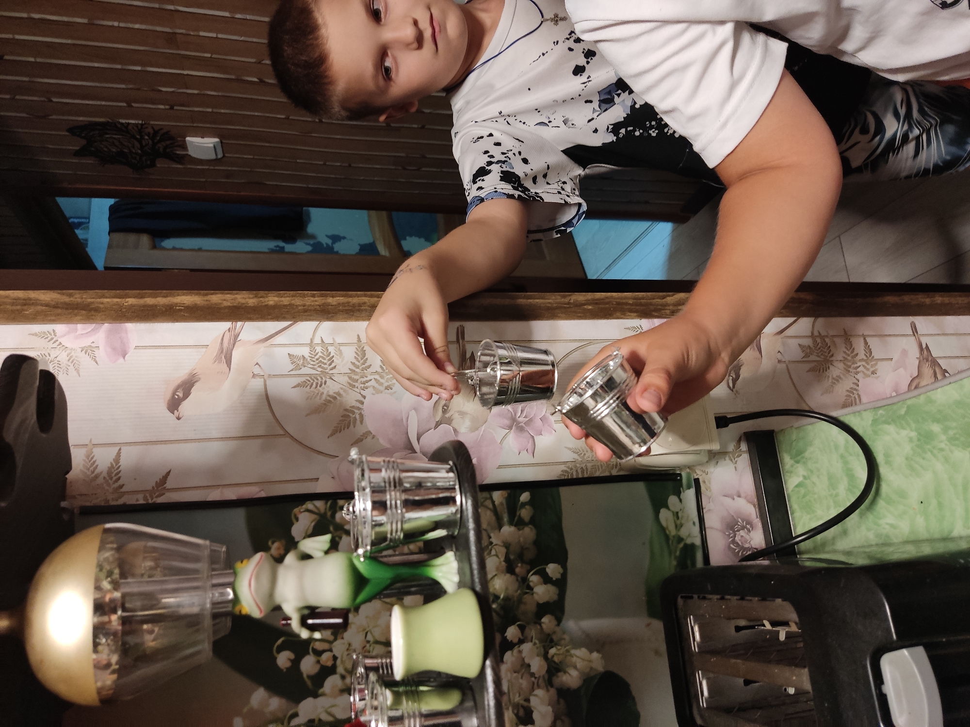 In a domestic interior setting, likely a kitchen or living area in Noginsk, Russia, a young boy with short brown hair and a white t-shirt with black splatters is holding a small, silver bucket. He is reaching out to place it onto a hook or a similar hanging mechanism, with another small silver bucket already attached. In the foreground, another similar silver bucket is held in his other hand. The background features floral wallpaper with a bird motif and a wooden paneled wall. A green ceramic frog figurine and several other small silver buckets are visible on a shelf. To the left, part of a black appliance, possibly a toaster, is visible. The lighting appears to be artificial.