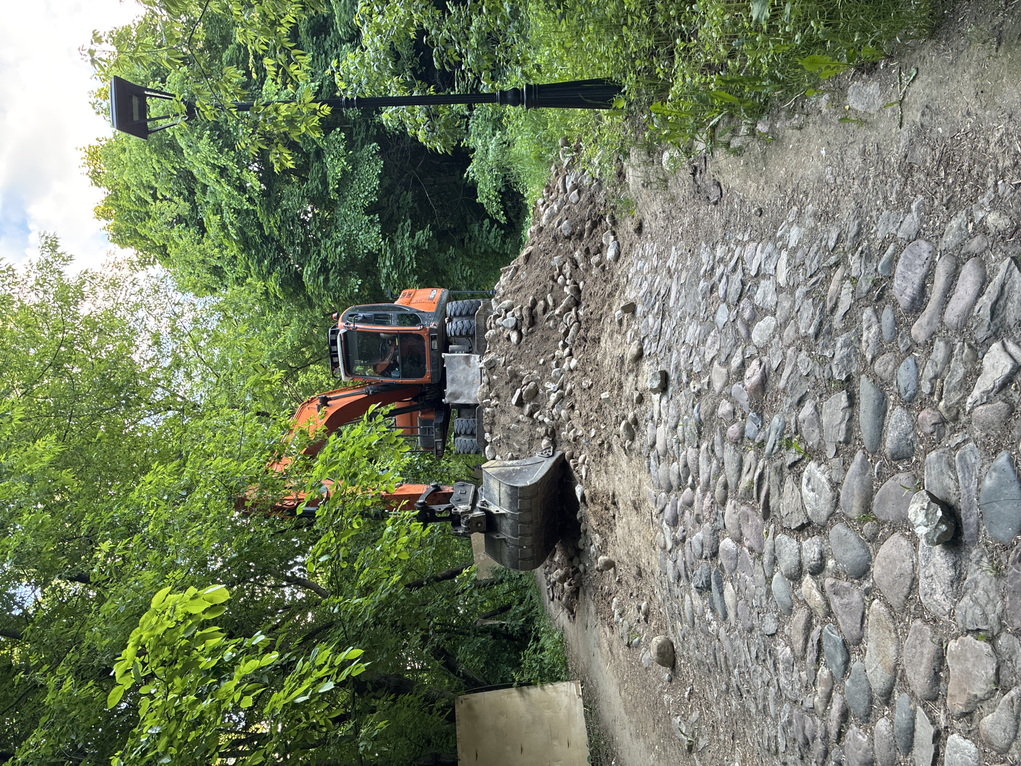 An orange excavator, likely engaged in maintenance work, is positioned next to a cobblestone path in a densely wooded area. A person is visible inside the excavator's cab. The machine's large metal bucket is near a substantial pile of dirt and rocks that extends alongside the path, suggesting it is actively involved in moving or clearing these materials.
The scene depicts an outdoor setting, likely a park or natural green space, given the abundance of lush green trees that frame the path and fill the background. A traditional black street lamp stands tall among the trees on the left side of the path. The path itself is constructed from old-style, irregularly shaped cobblestones, lending a rustic and possibly historical character to the area. A small, light-colored rectangular object, possibly a wooden board or container, lies on the ground near the excavator's bucket.
The time of day appears to be daytime, with bright, diffused light suggesting either an overcast sky or filtered sunlight. The vibrant green foliage indicates a warm season, likely spring or summer. The location is in Moscow, Russia. No visible text is clearly legible on any of the objects.