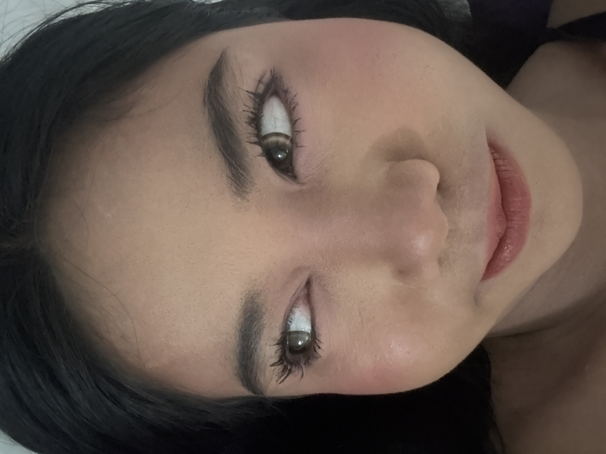 The image depicts a young adult female, likely of Asian descent, in an indoor setting within North Jakarta, Indonesia. Her dark hair is visible framing her face. She is wearing makeup, including foundation that provides an even skin tone, rosy blush on her cheeks, and dark mascara or false eyelashes that accentuate her eyes. Her eyes, featuring light brown irises, are directed towards the viewer's left. Her dark eyebrows are well-defined. Her lips are slightly parted, displaying a reddish-pink hue, and a subtle smirk suggests a coy or playful expression. The background is dark and undifferentiated, offering no specific environmental details beyond the subject.