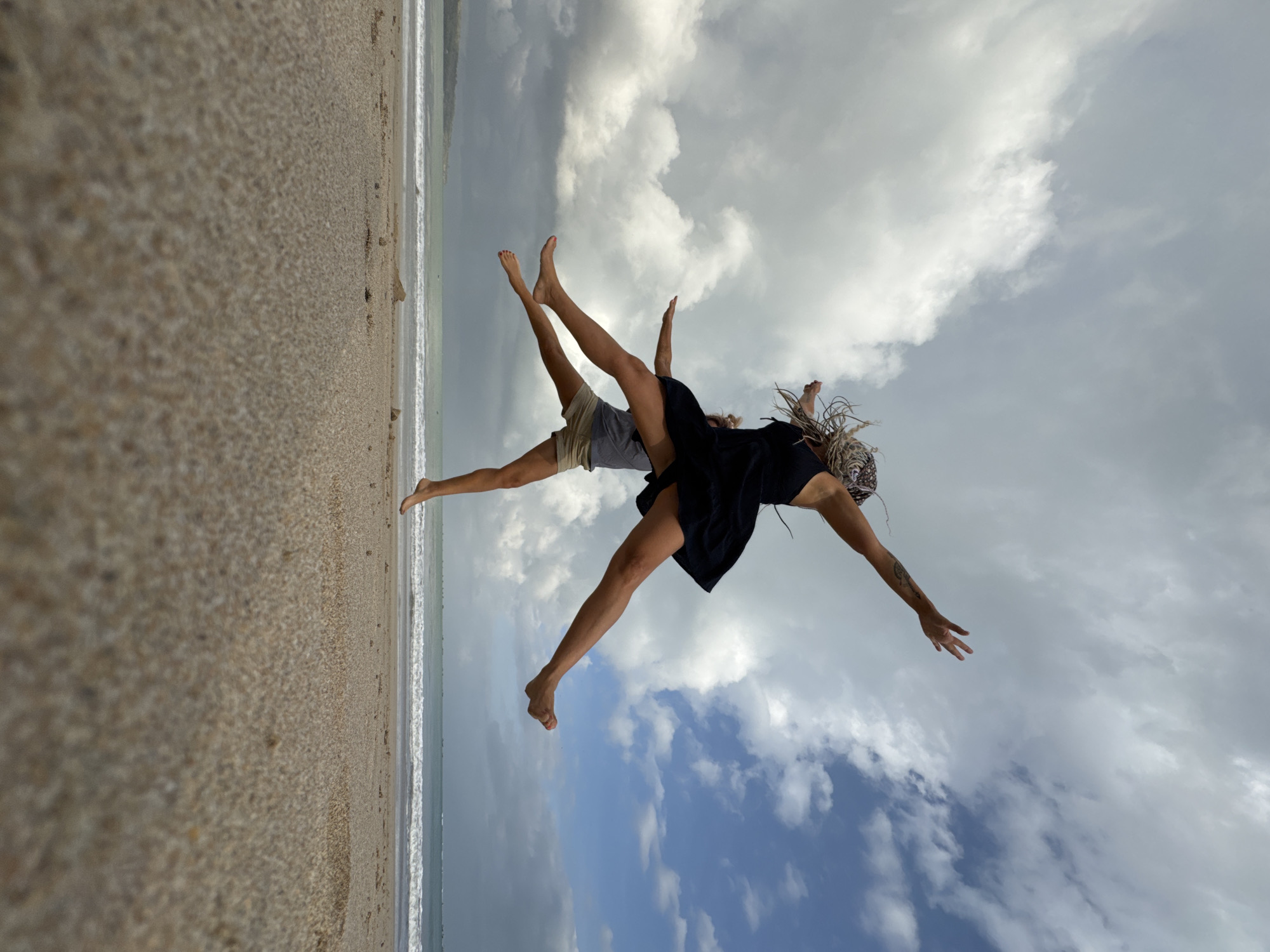 Two individuals are captured mid-air, jumping on a sandy beach with the ocean and cloudy sky in the background. The person in the foreground, closest to the camera, is positioned slightly below the other. They are wearing tan shorts and a grey t-shirt, with their legs extended outwards and slightly bent. The second person, positioned above and behind the first, is wearing a black dress and has their legs spread wide. Their arms are outstretched, and their hair is styled in blonde dreadlocks. The scene appears to be on a beach in Kuta, Indonesia, characterized by its sandy shore, gentle waves, and a distant shoreline. The lighting suggests it is daytime, with a mix of sunlight and cloud cover contributing to a dynamic sky. The perspective is low, emphasizing the upward movement of the individuals.