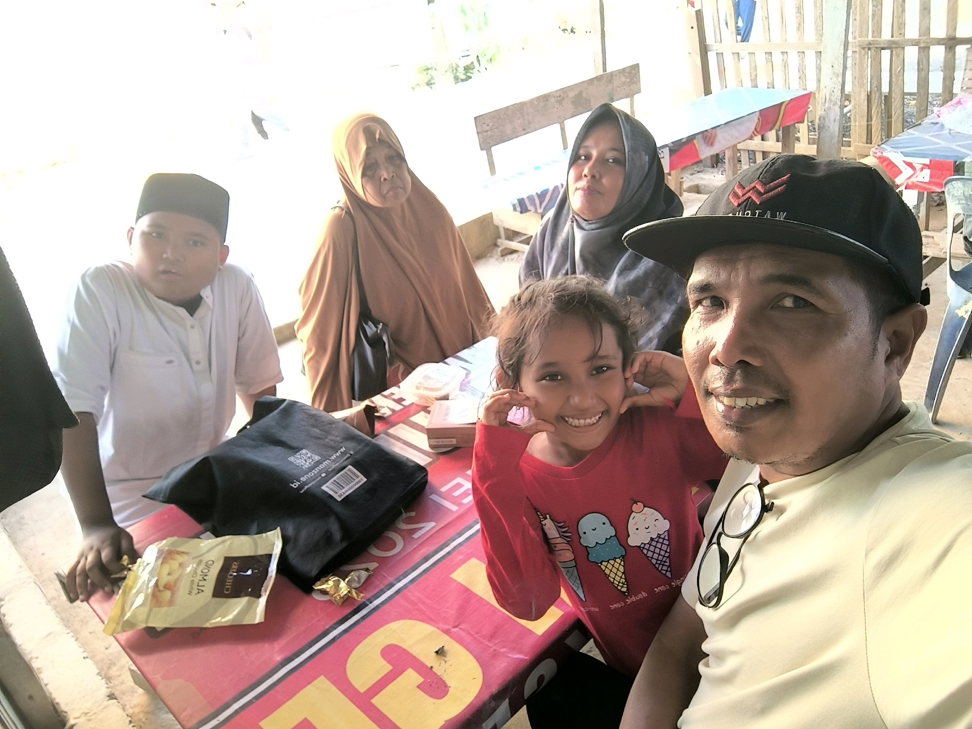 A group of five individuals, two adult males, two adult females, and one young girl, are captured in a selfie-style photograph, seated around a pink table. The individuals appear to be of Malay ethnicity. The setting suggests an outdoor, casual environment, possibly a roadside eatery or a gathering in a public space in Lhokseumawe, Indonesia. The background reveals wooden structures and what appears to be a beach or coastal area, indicated by the presence of sand and a body of water. The people are dressed in casual attire, with the younger individuals wearing a white shirt and a red shirt respectively, and the adults in traditional Islamic head coverings and t-shirts. The table is adorned with various items, including snacks, a black bag, and a pair of glasses. The overall mood is light and familial, with the child in the foreground smiling and posing for the camera. The image quality is moderate, with some overexposure in the background, suggesting it was taken during daylight hours.
