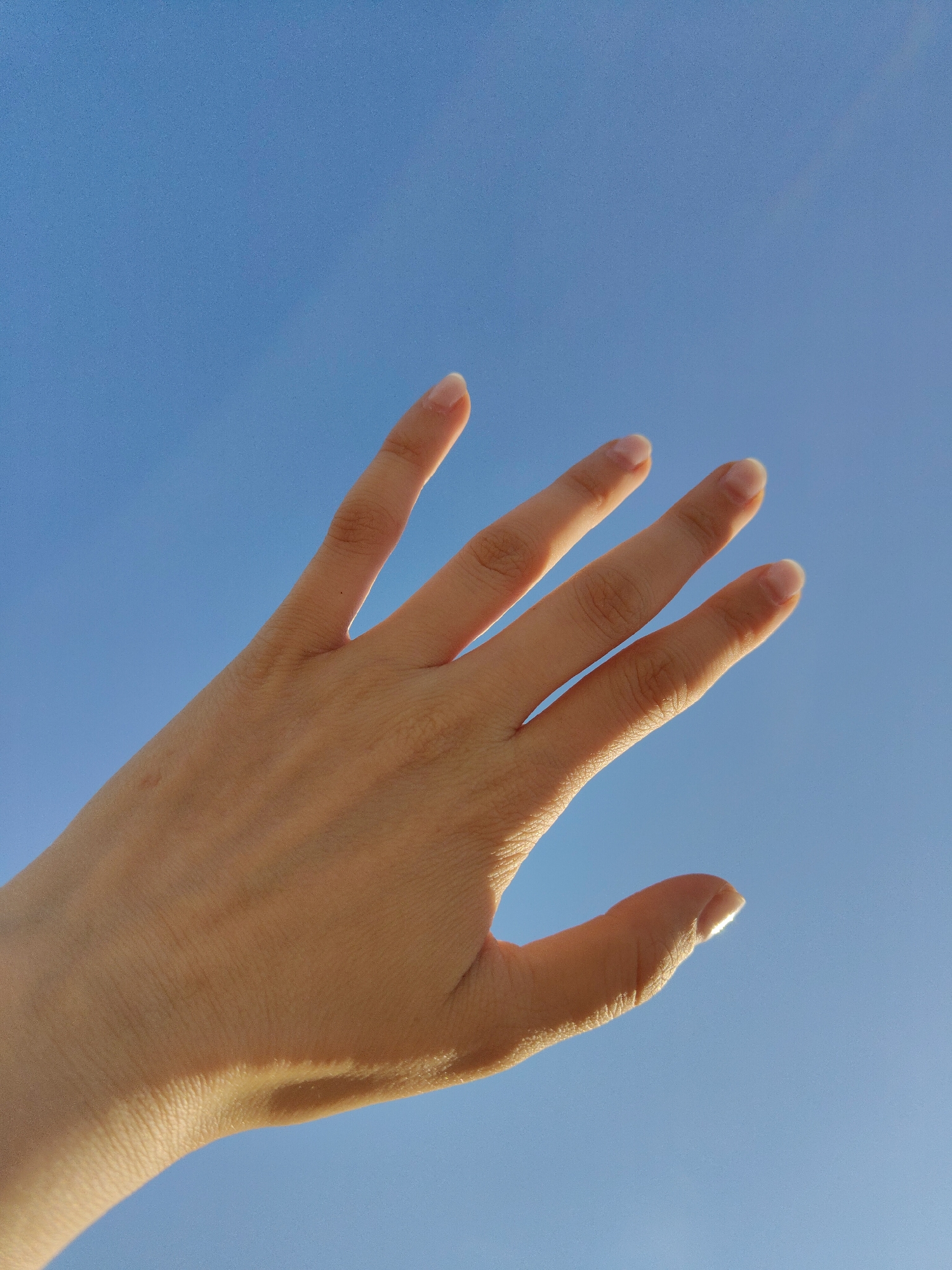 A right hand is held up against a clear blue sky. The fingers are spread slightly apart. The hand is in focus, with the blue sky as a backdrop.