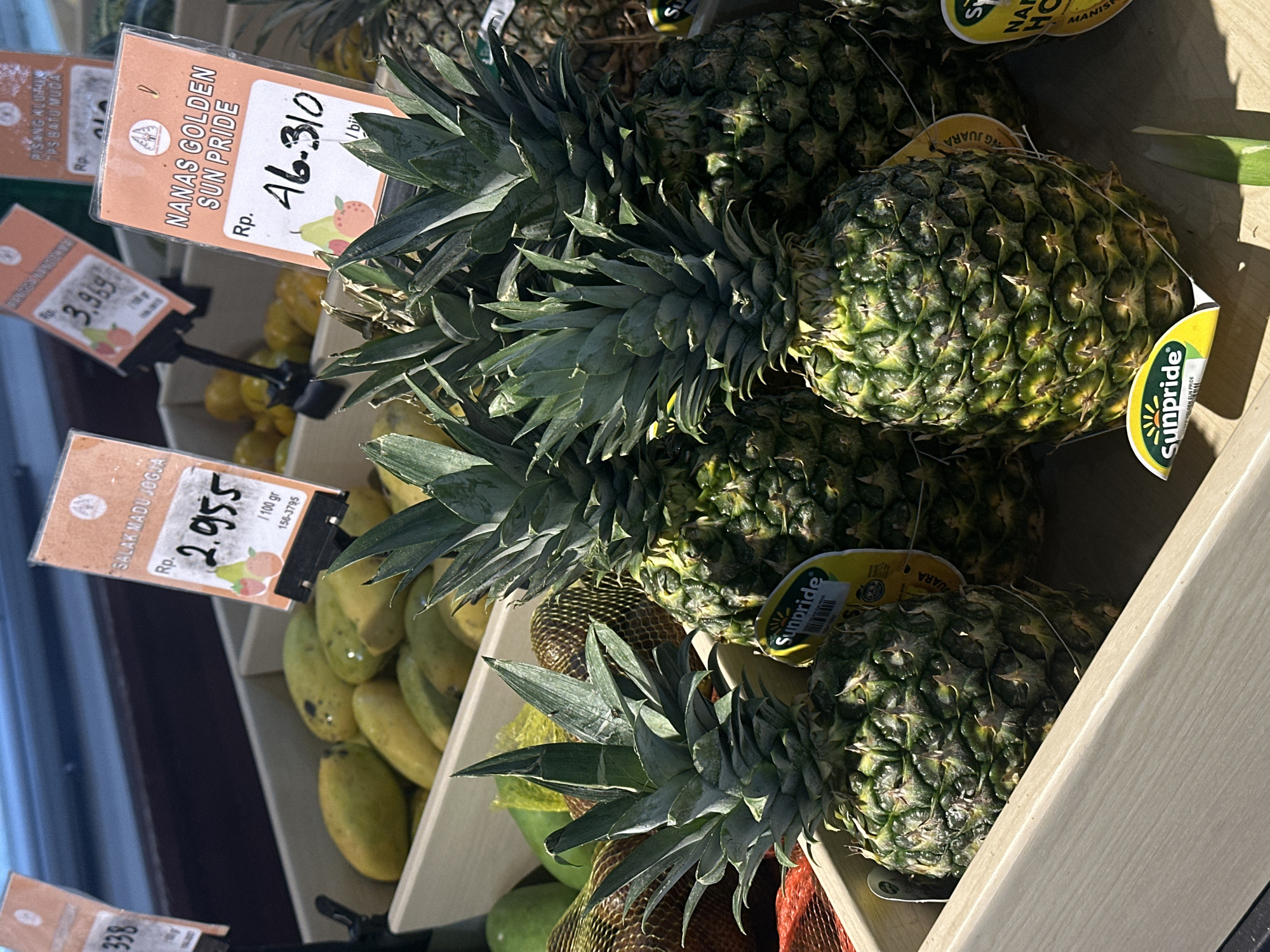 This is a close-up, eye-level shot of a produce display in a market, likely a grocery store, in North Kuta, Indonesia. The focus is on several ripe pineapples in the foreground, arranged in a light-colored wooden crate. The pineapples have rough, spiky green and yellow skin and leafy green crowns. Yellow stickers with the brand name "Sunpride" and some text, partially obscured, are attached to the pineapples. Behind the pineapples, another crate is filled with light green and yellow mangoes. In the background, more produce is visible, including what appear to be more mangoes and possibly other fruits, all bathed in bright, natural light. Several price tags are visible, indicating prices in Indonesian Rupiah (Rp). One tag reads "SALAK MADU JOGJA" with a price of "2.955". Another tag above the pineapples reads "NANAS GOLDEN SUN PRIDE" with a price of "Rp. 46.310". The overall impression is of a fresh produce section within a retail environment.