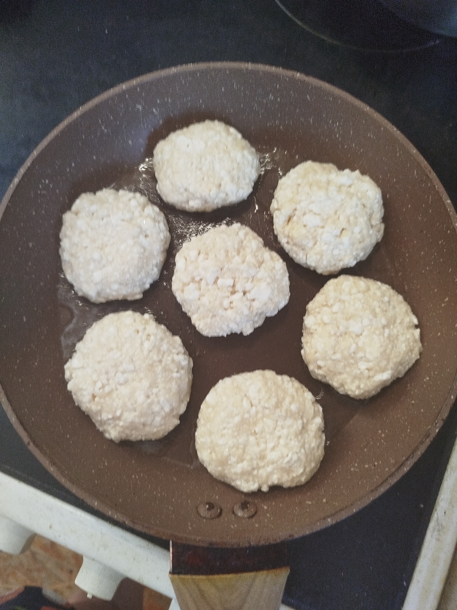 Seven round, pale-yellow patties, possibly potato pancakes or fritters, are arranged in a circle within a dark brown, non-stick frying pan. The patties have a slightly uneven, textured surface. The pan itself appears to be on a stovetop, with visible elements of a white appliance below, and a dark countertop behind. There are no people or animals present. The scene is indoors, likely a kitchen. The image does not provide any specific information to indicate its location in Zavodoukovsk, Russia.