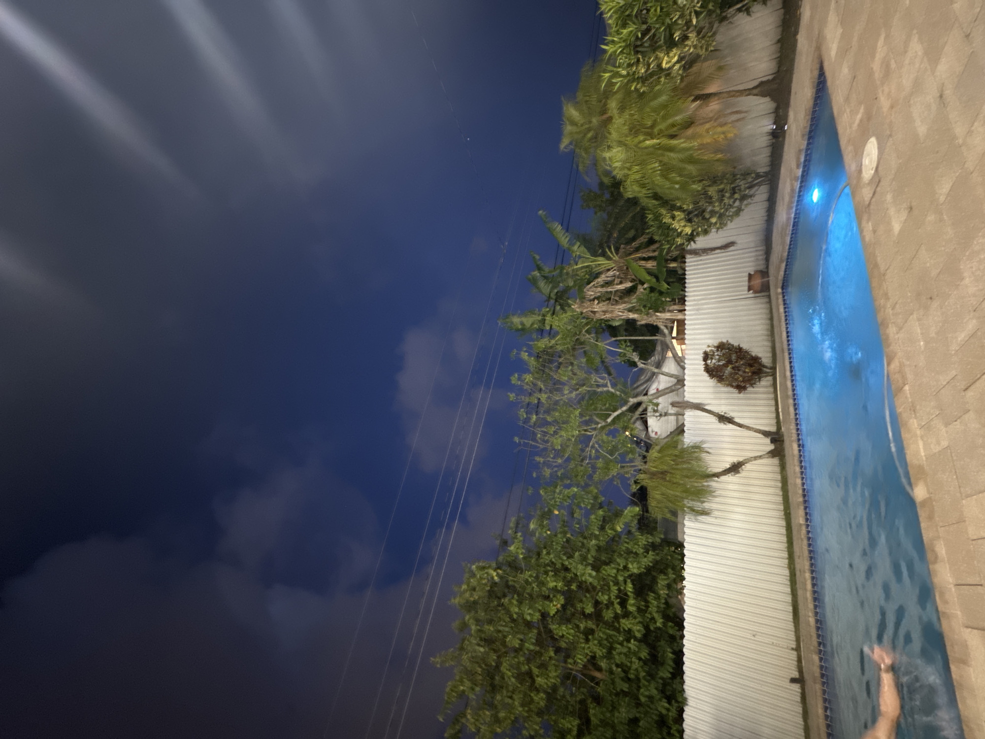 The image depicts a nighttime scene in what appears to be a residential backyard in Miami, United States. The scene is oriented vertically, with the dark blue sky and scattered clouds occupying the left half of the frame, contrasted by illuminated plants and a swimming pool on the right half. Utility lines are visible against the sky.
The backyard features a row of trees and plants along a white fence that borders the pool. The pool itself is brightly lit with blue light, and its water surface shows slight ripples and movement. A person’s arm extends upward from the lower right corner of the pool, suggesting someone is swimming or standing in the water. The pool's edge is lined with light-colored pavers. The overall environment suggests a warm, tropical climate, consistent with Miami's geographic location.