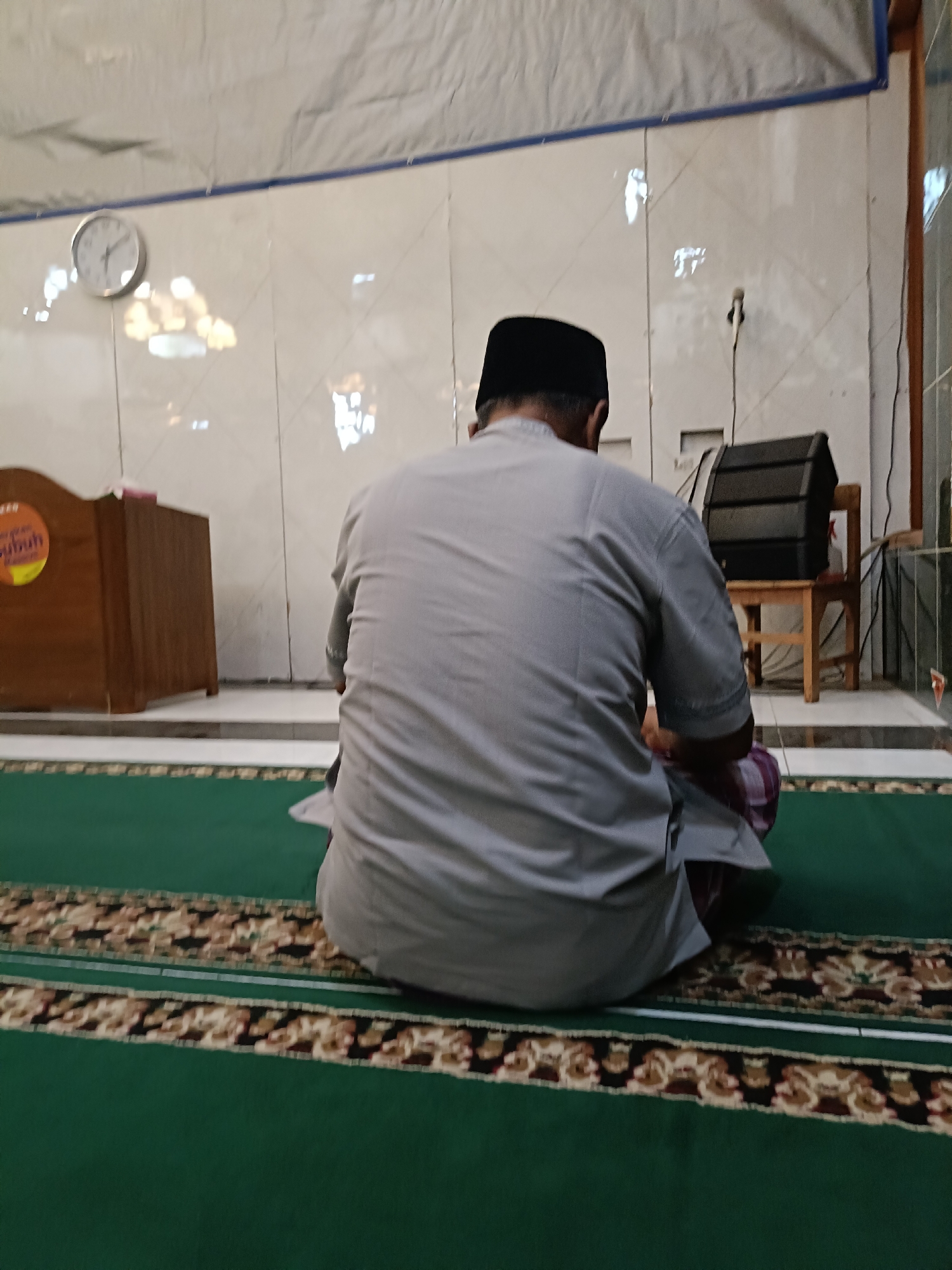 A man in a grey kurta and black peci cap is seen from behind, sitting on a green prayer rug with a patterned border. He appears to be praying or meditating in a mosque or prayer hall, indicated by the presence of a wooden pulpit to his left, a clock on the wall behind him, and a black speaker on a wooden chair to his right. The walls are tiled and there is a white ceiling with some lighting fixtures. The floor is covered with a green carpet and a patterned runner. The image appears to be taken indoors, possibly in Kediri City, Indonesia.
